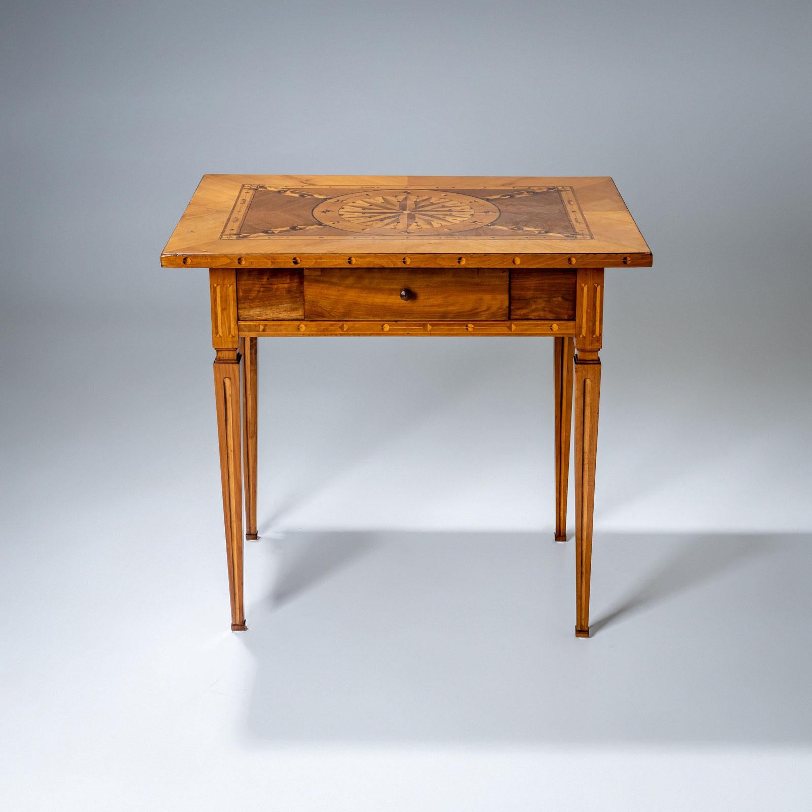 Table d'appoint à plateau rectangulaire et hauts pieds fuselés. La marqueterie du plateau de la table forme un médaillon central avec rosace et se poursuit avec des éléments décoratifs de marque tels que des cannelures et des perles implicites. Le