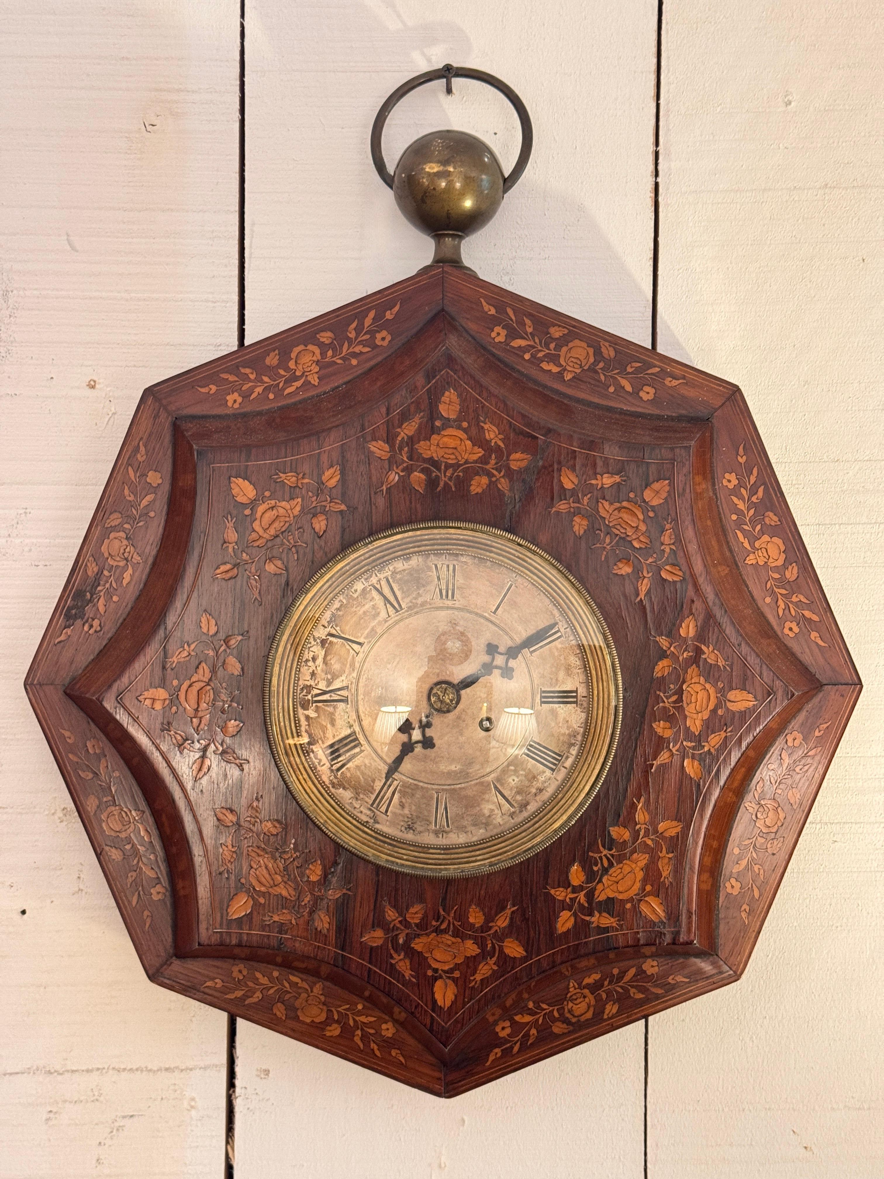 Decorative wall clock with shaped octagonal form and fine marquetry inlay of vines and roses throughout. The silvered dial with Roman numerals shows notable age and patina. A round brass finial and loop completes the top. The detailed floral inlay