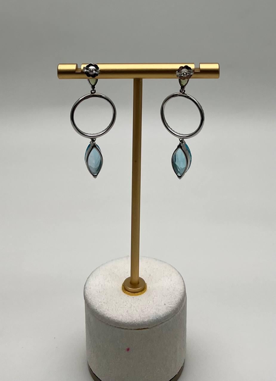 These contemporary earrings showcase a striking design with circular diamond-studded hoops. Each earring features a marquise-cut blue gemstone topaz, complemented by a smaller green peridot at the top. 14K White Gold. 0.40 CTW Diamonds. 7.83 CTW