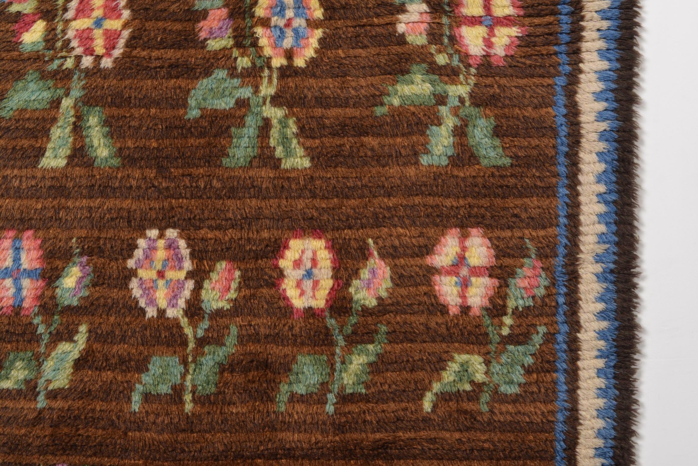 Created by Swedish textile designer Märta Måås-Fjetterström in the early to mid 20th century, this traditional wool hand-woven Swedish pile rug has features a rich brown base with multi colored vivid floral patterns and a blue and cream boarder.