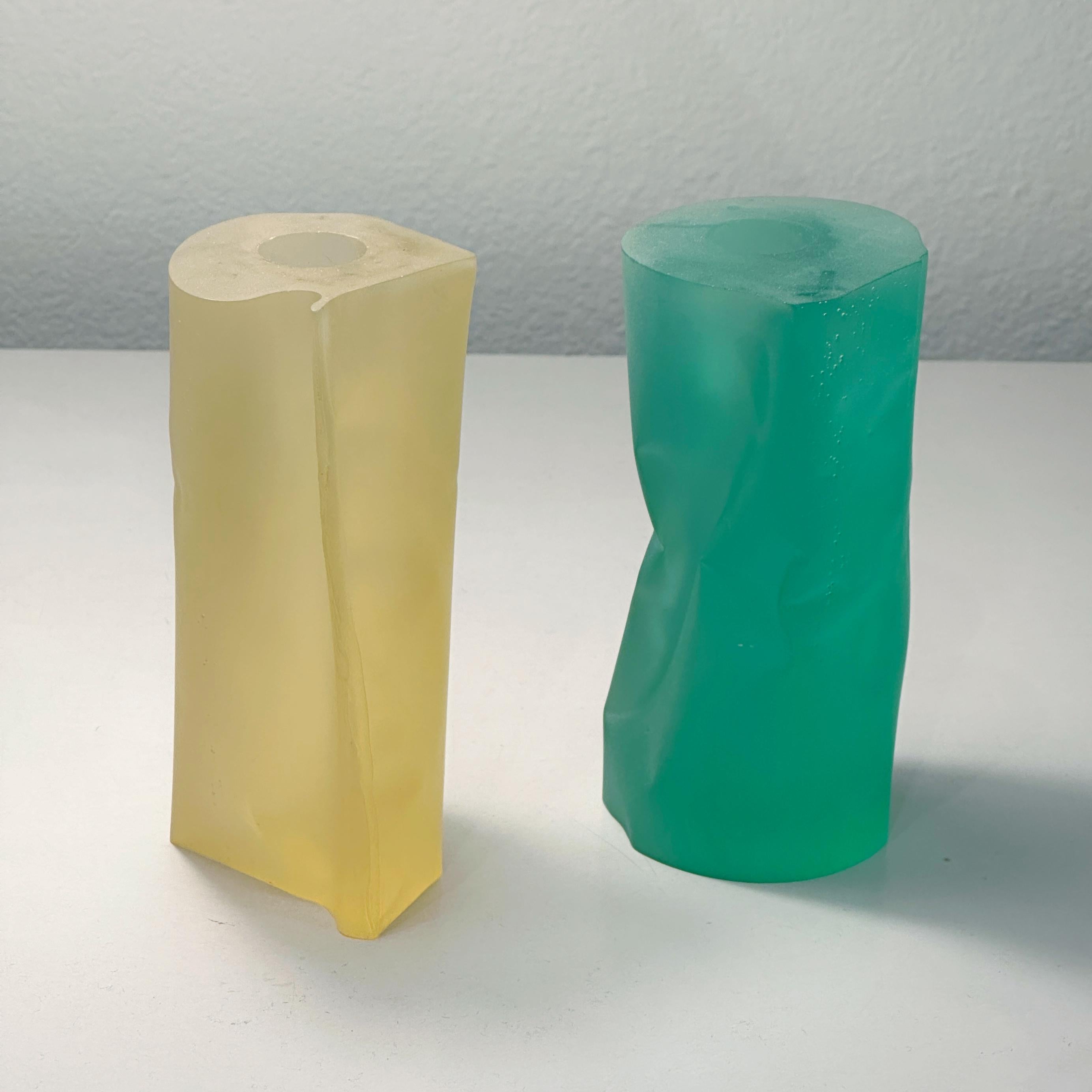 A pair of opaque resin candle holders, one green and the other a light yellow resin.  The cylindrical shapes are asymmetrical, solid and confident but have been carved as if they were paper or metal cylinders having been lightly crushed. One candle