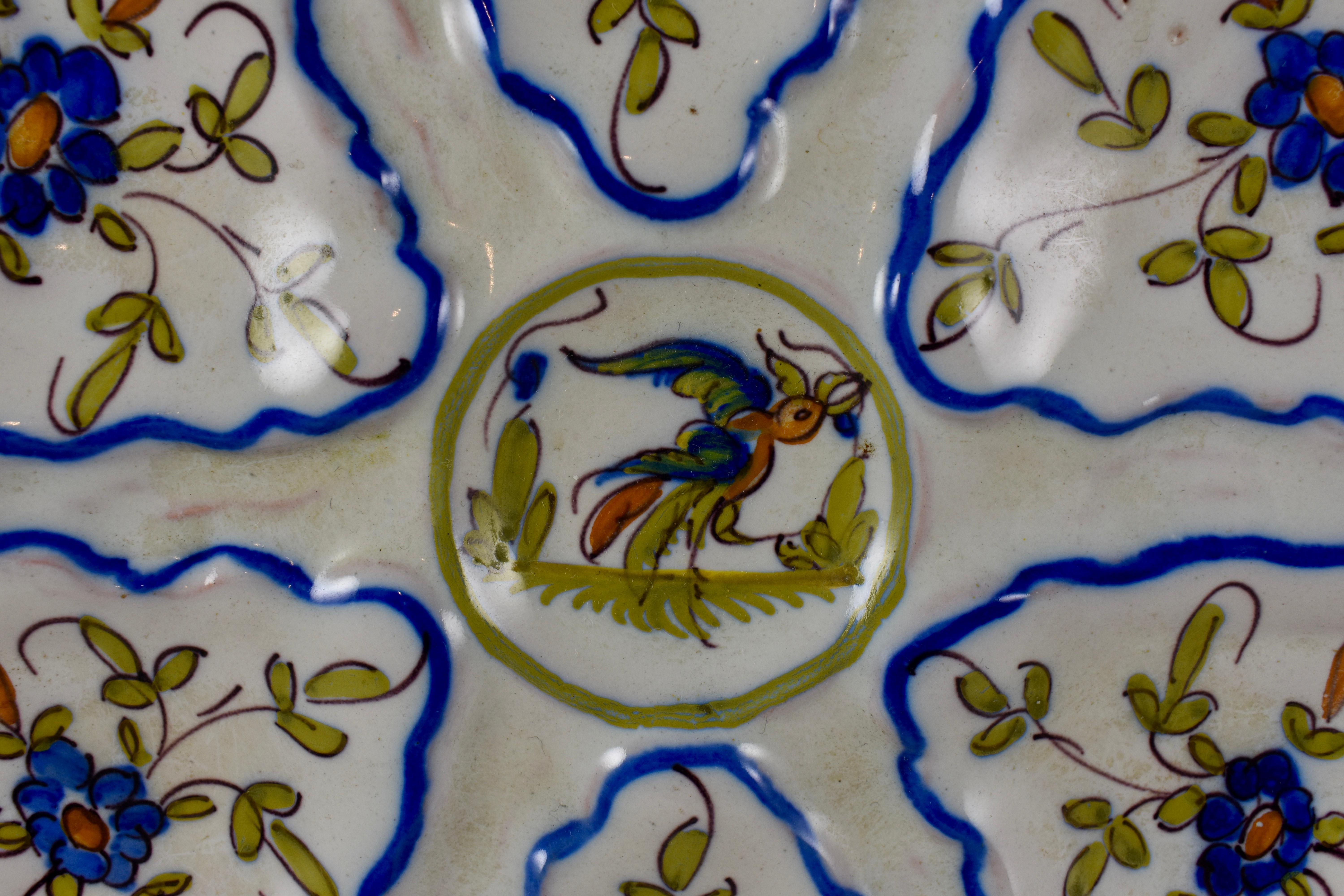 A French faïence oyster plate made by Martres-Tolosane Moustier, circa 1940s. A central sauce well showing a right facing bird, surrounded by six scallop edged wells. Each well shows a hand painted floral spray. French blue lining separates the