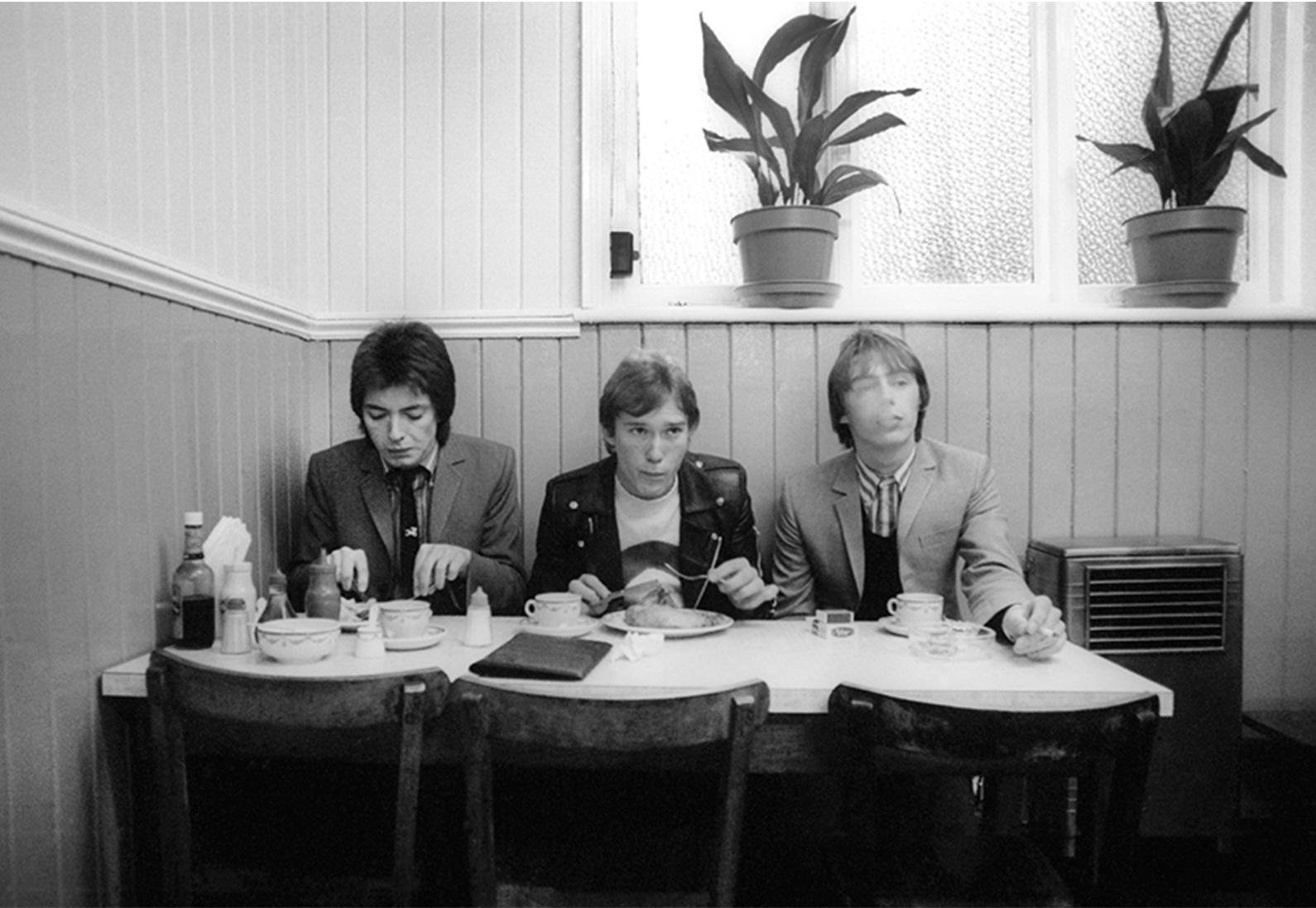 The Jam entspannen sich in Frank's Cafe in der Lexington Street, Soho, während ihres Fotoshootings für die Titelseite von 'News of the World'. Aufgenommen vom Fotografen Martyn Goddard im Jahr 1978.

Martyn Goddard erinnert sich: "Für das Cover von