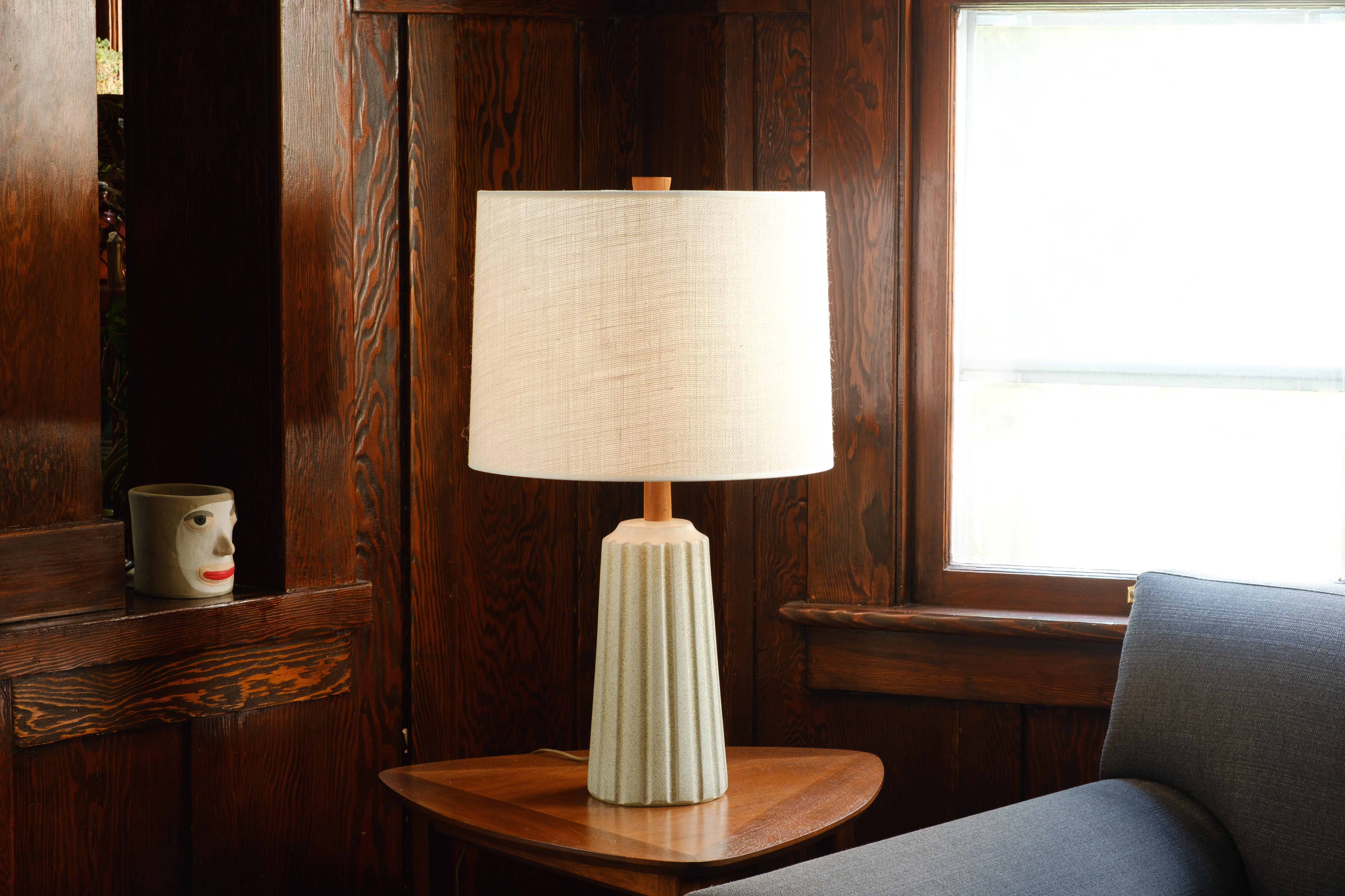What is it?
—
This unusual Martz lamp comes in a matte cream glaze with embedded bits of sand. Reminiscent of a column, indentations surround the perimeter. The ceramic body gives way to a walnut neck and the whole ensemble is capped off with an