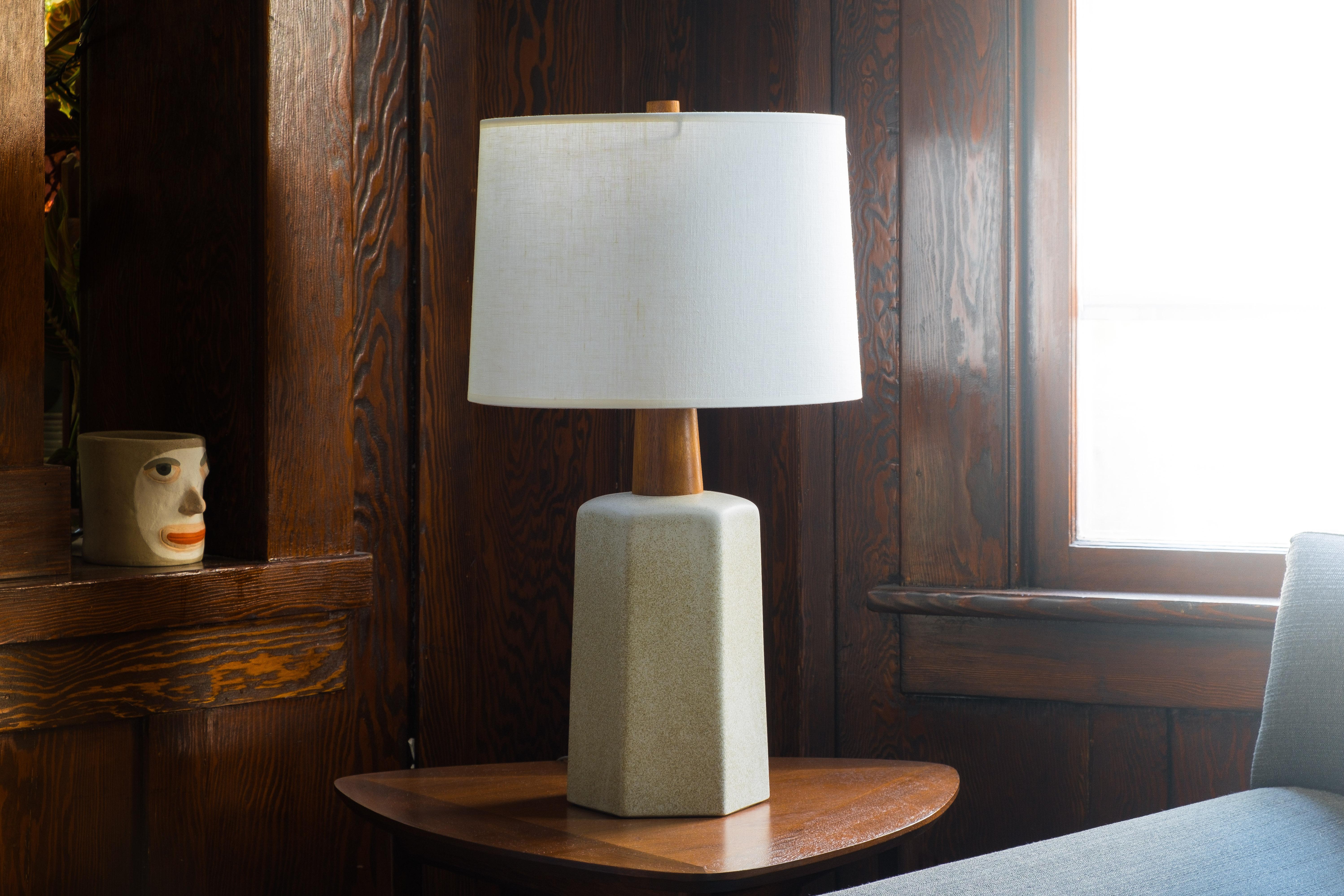 What is it?
—
This unusual Martz lamp comes in a matte cream glaze with embedded bits of sand. The body is a tapered hexagon shape with a top that rounds gently. The ceramic body gives way to a thick walnut neck and the whole ensemble is capped