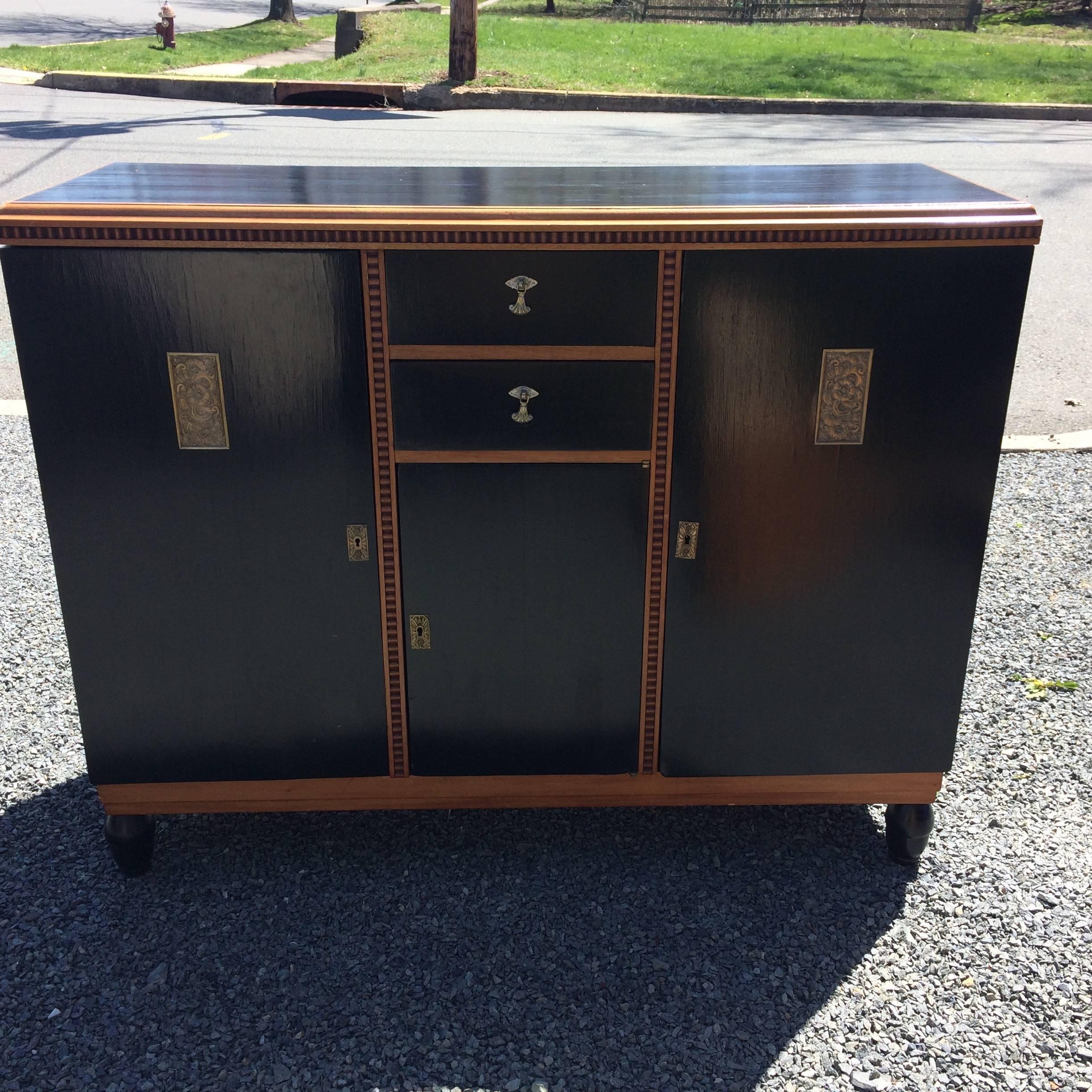 A rare vintage Art Deco style ebonized wood cabinet having contrasting carved wood detailing an charming embossed brass decoration and hardware. Two small drawers and three panel doors reveal storage inside. Has a key that opens the doors, but