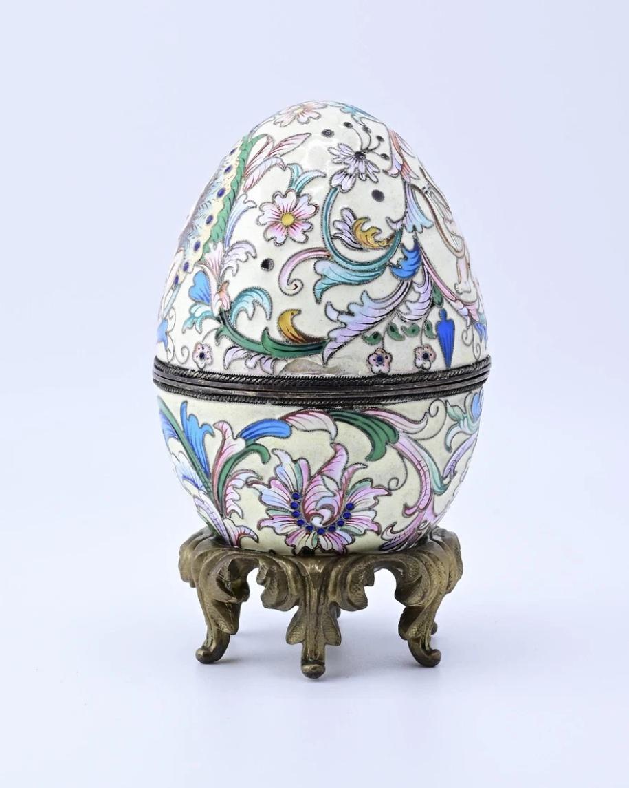 Esta exquisita huevera rusa decorativa de plata esmaltada es una pieza cautivadora. Adornada con intrincados motivos florales en suaves tonos pastel, irradia elegancia y encanto. El huevo está fabricado con meticulosa atención al detalle, y presenta