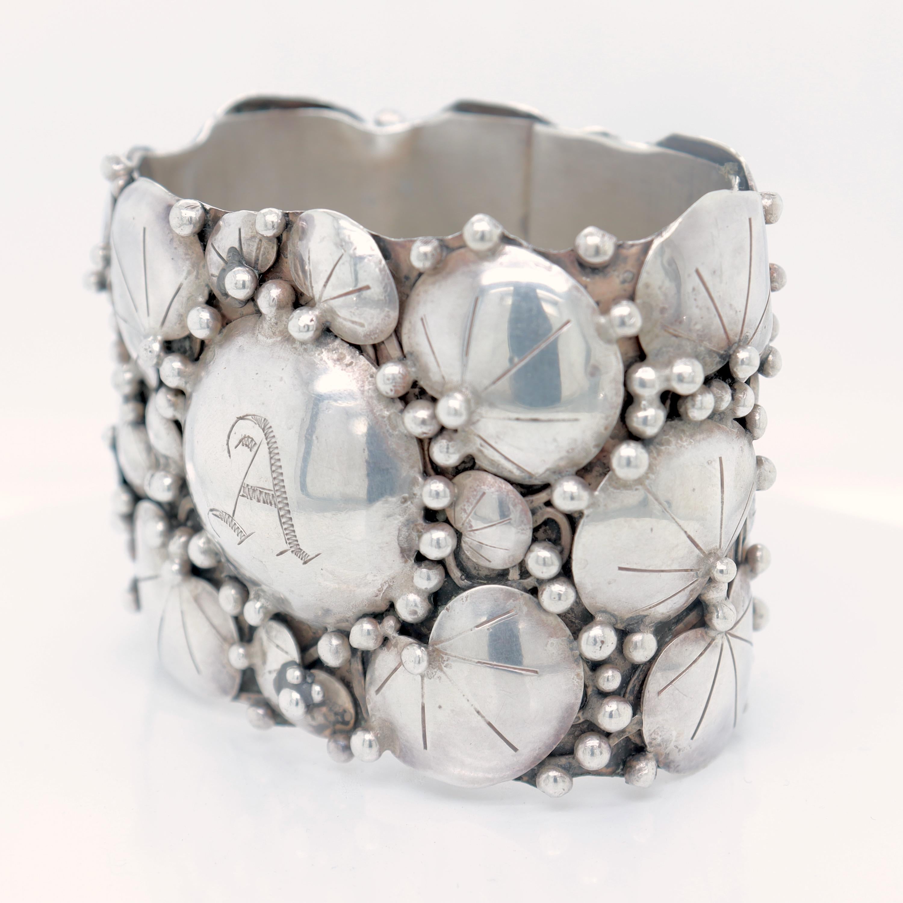 A wonderful Arts and Crafts napkin ring.

By Mary Gage, the renowned early 20th Century American female silversmith.

In sterling silver with stylized lil pads surrounding a central raised button cartouche with a monogrammed 