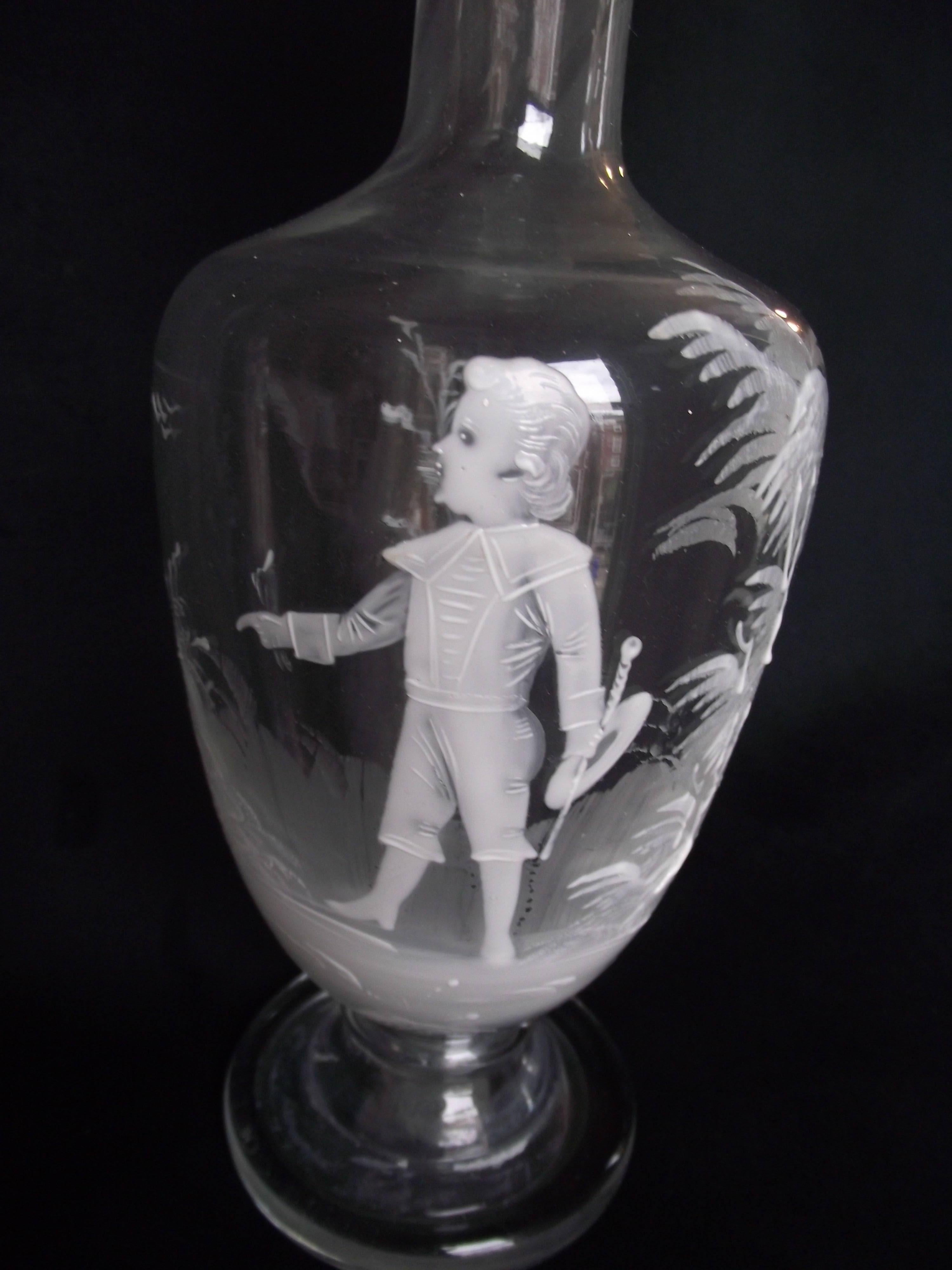 This mouth blown decanter is hand painted by Mary Gregory in white enamel. It features an intricate painting of a young boy holding a plant stem in one hand and his hat and stick in the other. The surroundings are tropical with palms and ferns,
