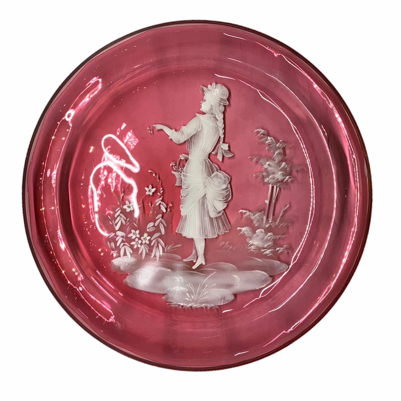 Il s'agit d'une assiette en verre émaillé blanc de Mary Gregory de couleur canneberge claire représentant une scène d'une élégante jeune fille de l'époque victorienne cueillant des fleurs dans un jardin et les mettant dans son panier.