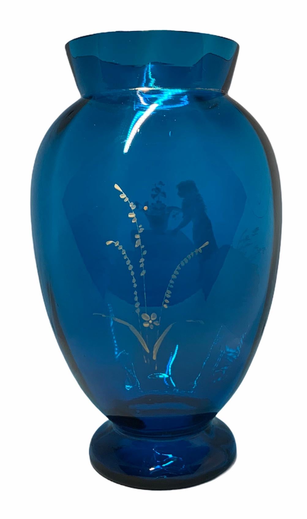 This is an urn shaped royal blue glass vase depicting a white enamel painting of a young girl standing in front of a table while making an arrangement of flowers.