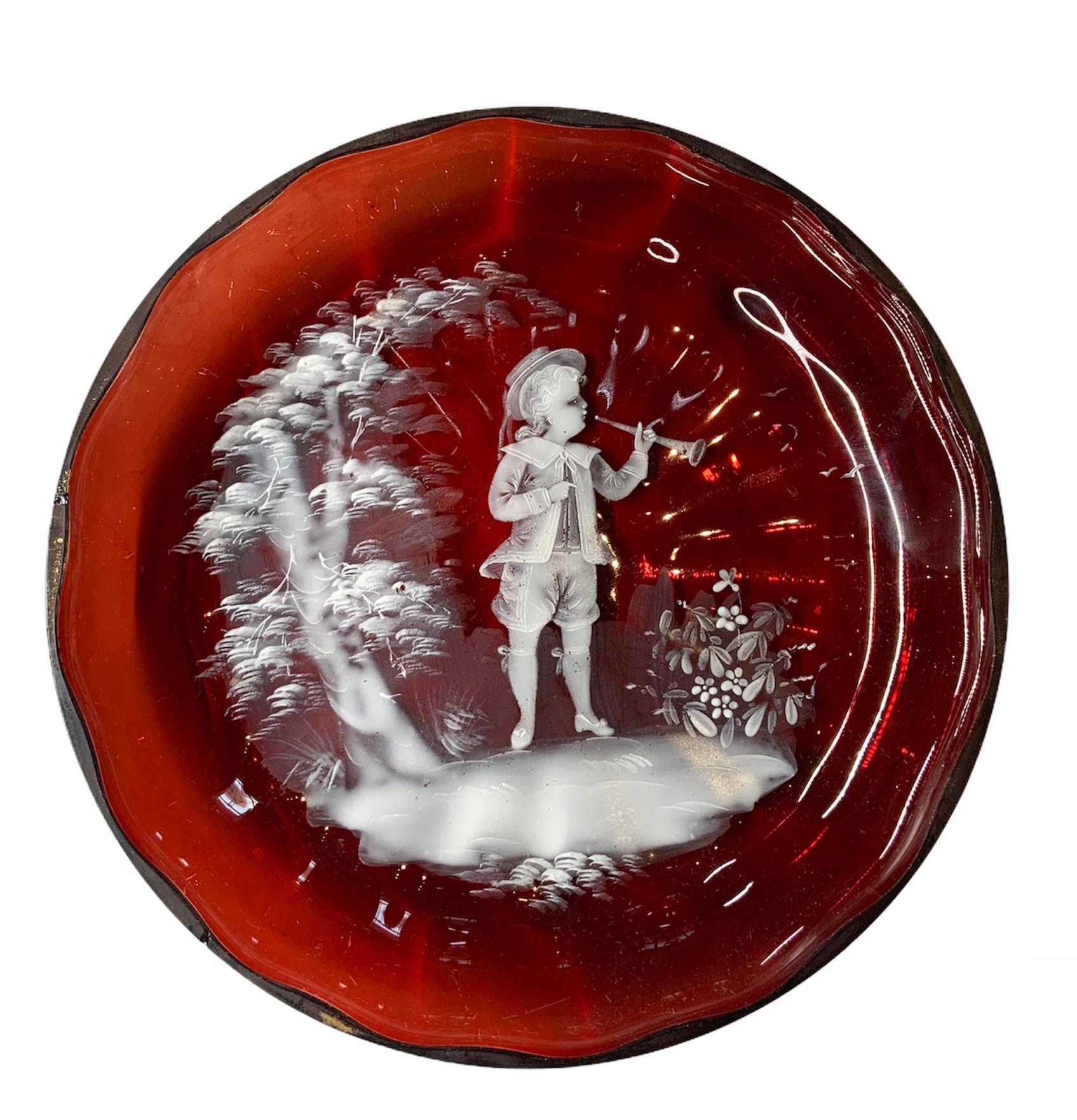 This a Mary Gregory ruby red white enameled glass featuring a scene of a Victorian era boy playing a trumpet in a landscape adorned with a small tree and some flowers.
