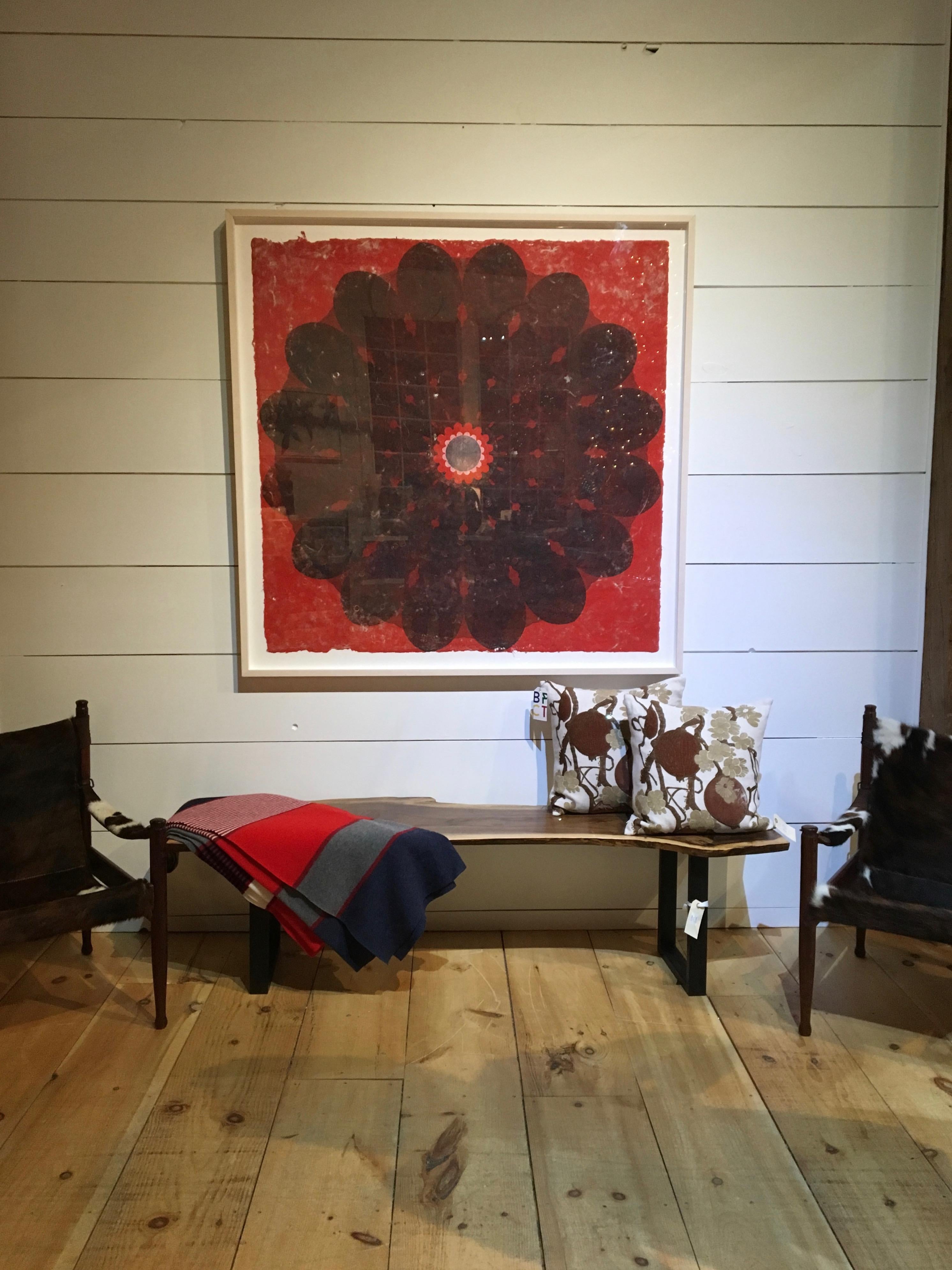In this large, square relief print on handmade paper, a circular, botanical mandala shape with petal-like edges in dark grey is dramatic and eye-catching against the brilliant red background. A circle surrounded by smaller petal shapes is at the