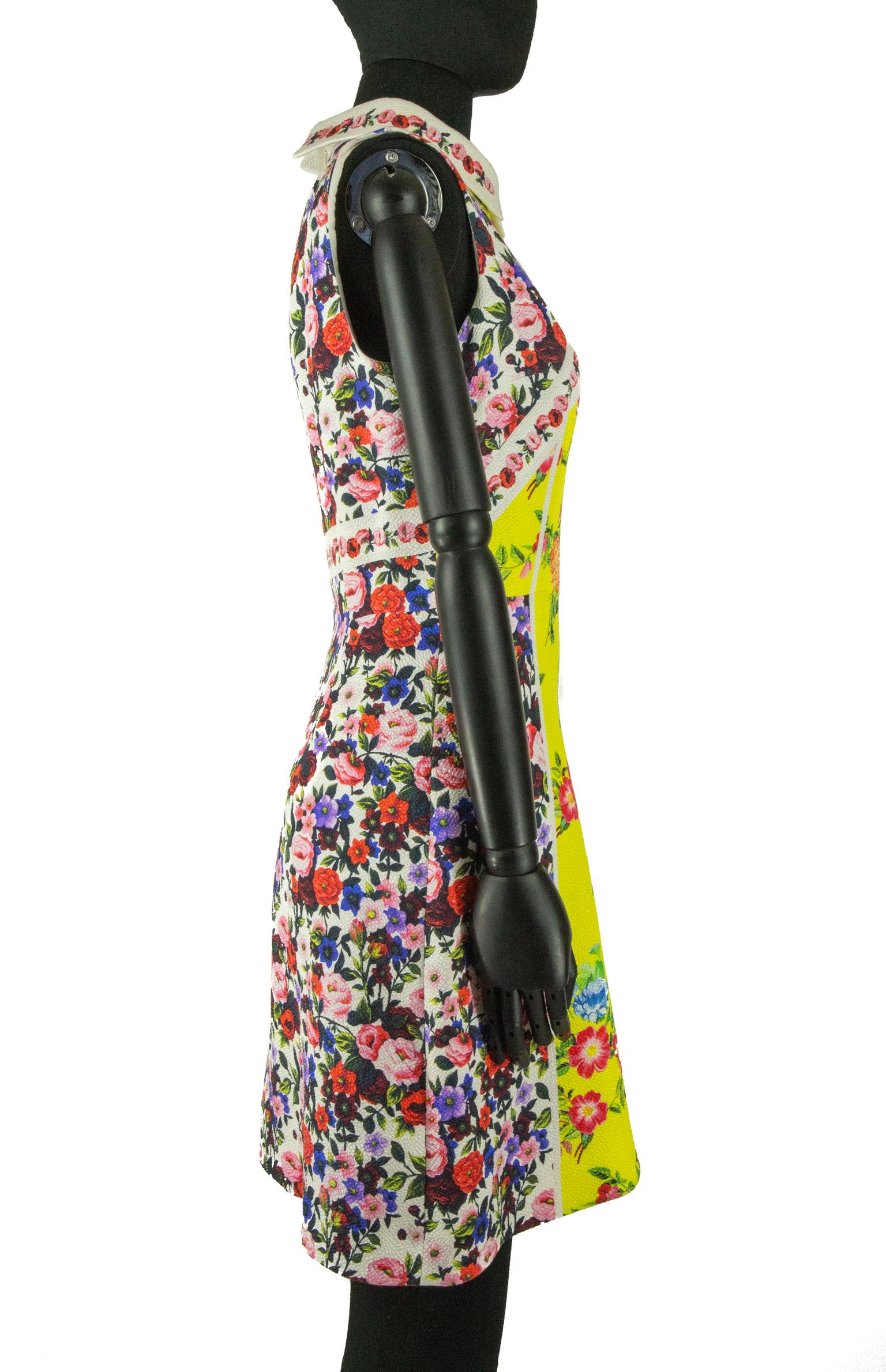 A Mary Katrantzou skater dress adorned in the brand's iconic prints, of symmetrical floral print with roses and peonies in shades of pink and red on a lemon yellow. The darted bodice is sleeveless with a standard collar, attached to an A-line skirt.
