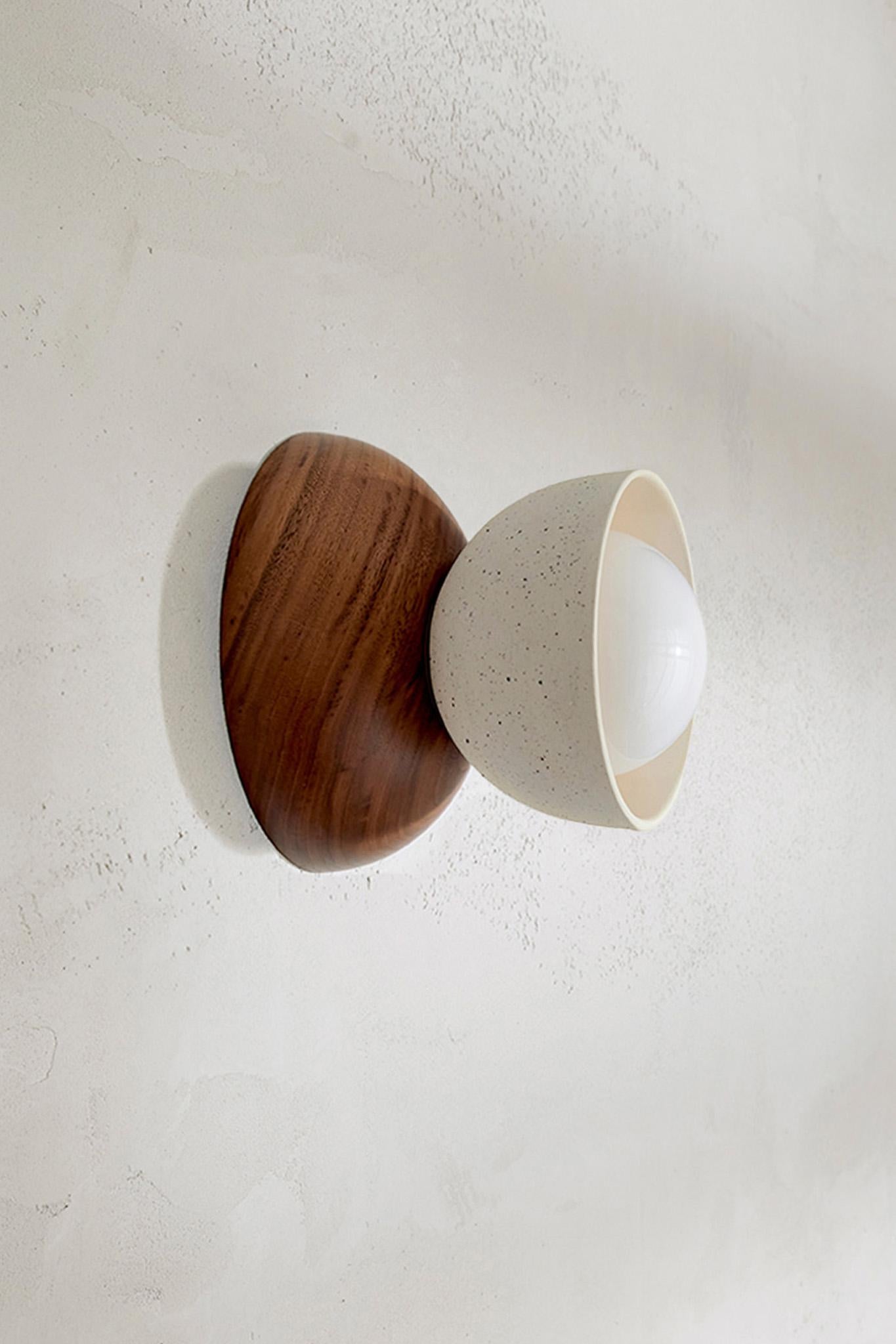 Die Terra 00 Surface Sconce besteht aus einer Kombination von zwei kontrastierenden Halbkuppeln, einem Schirm aus gegossener Keramik und handgedrechselten FSC-zertifizierten Hölzern. So entsteht eine moderne Form, die zwei Elemente miteinander