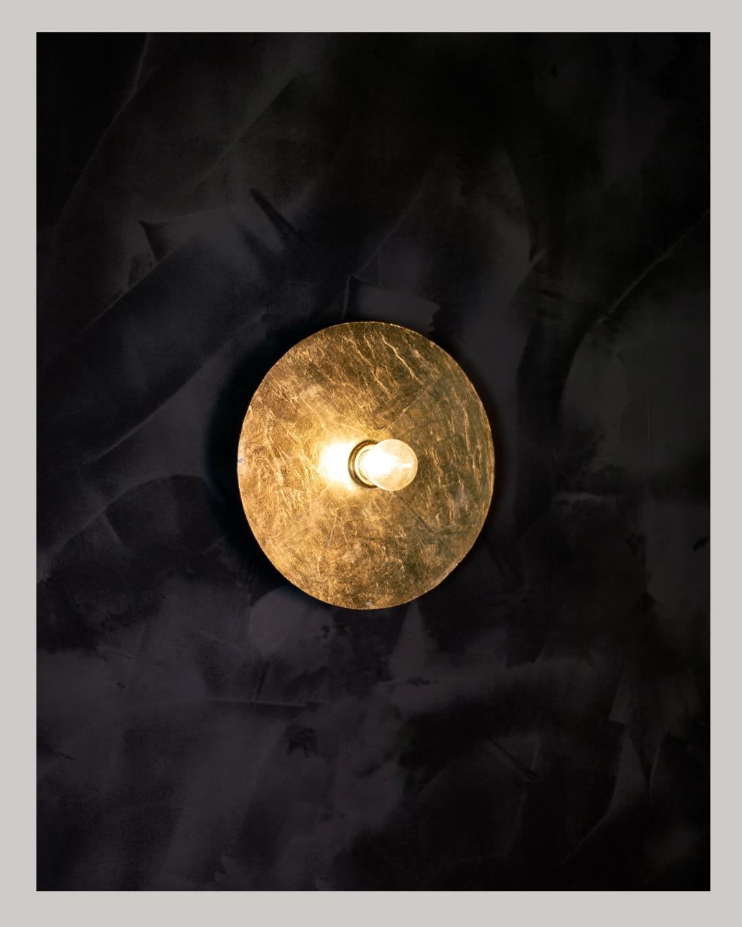A metal disc hand gilded with gold leaf. Each fixture is unique in it's finishing. Suitable for either wall or ceiling applications. The fixture casts a very unique shadow while emitting a warm and comforting light from the reflection of the gold