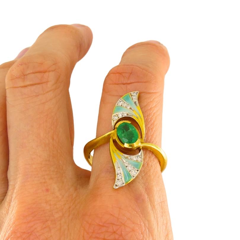 A beautiful Masriera Art Nouveau style ring, made of 18k gold, featuring emerald, diamonds, and enamel.
The ring’s design has a flowing, asymmetrical feel. Two wing like shapes extending from the center to curve around the deep green emerald. 
The