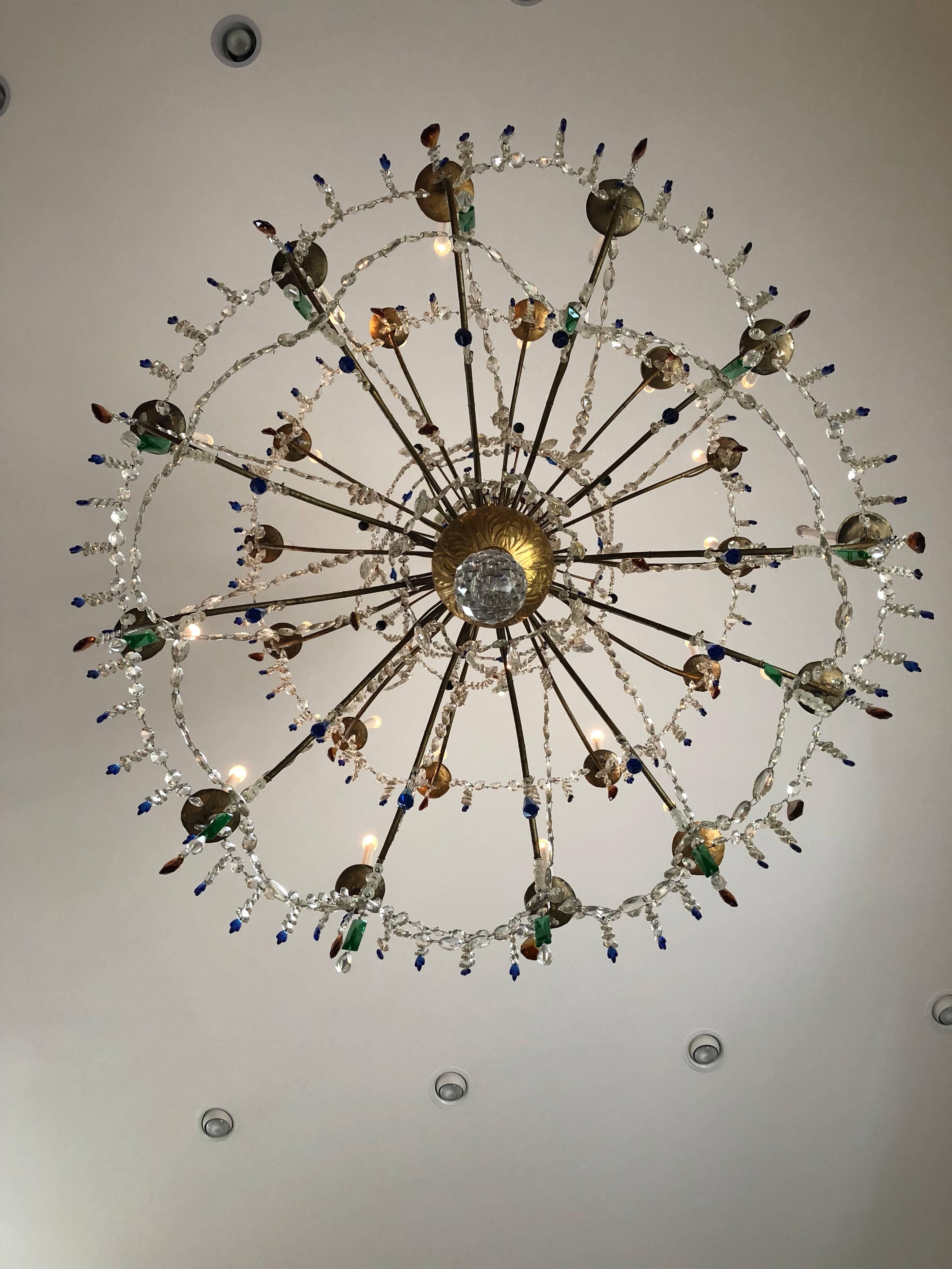 A massive 19th century Italian Genoese giltwood and crystal chandelier. With carved giltwood centre with two tiers of twelve lights draped with clear, emerald, amber, and royal blue crystals.