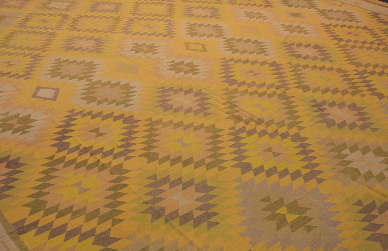 This is a massive antique Indian Dhurrie Rug flat-woven circa 1920 - 1950 and measure 620 x 405CM in size. It has an overall design with repeating sawtooth medallions set on a saffron yellow background to color. Its main border has an