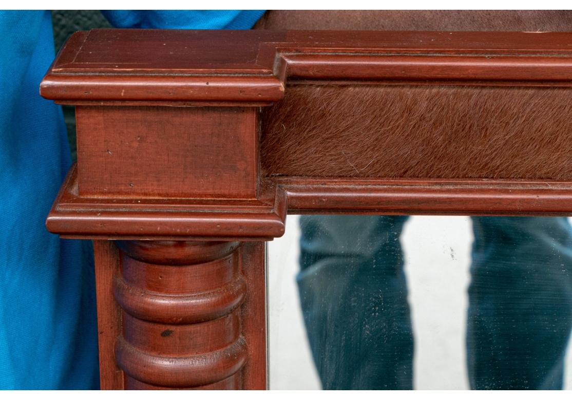 Un grand miroir rectangulaire solide avec un cadre en bois de cerisier avec des accents de peau de vache en haut et en bas. Le cadre présente un motif strié répétitif le long des côtés et des sections lisses recouvertes de peau de vache en haut et