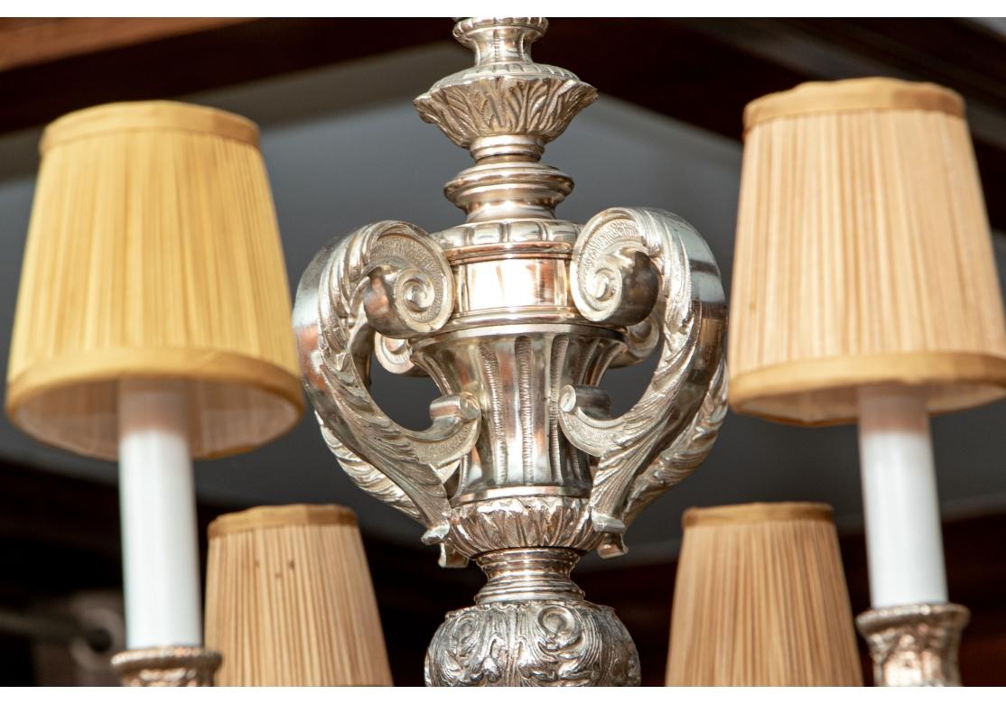 A silver finished metal Classic Baroque style chandelier with ten leafy scrolled lights covered with tan pleated shades. The standard with four scrolls on the top of a shaped center. A large scale bulbous terminal with artichoke end. All bobeches