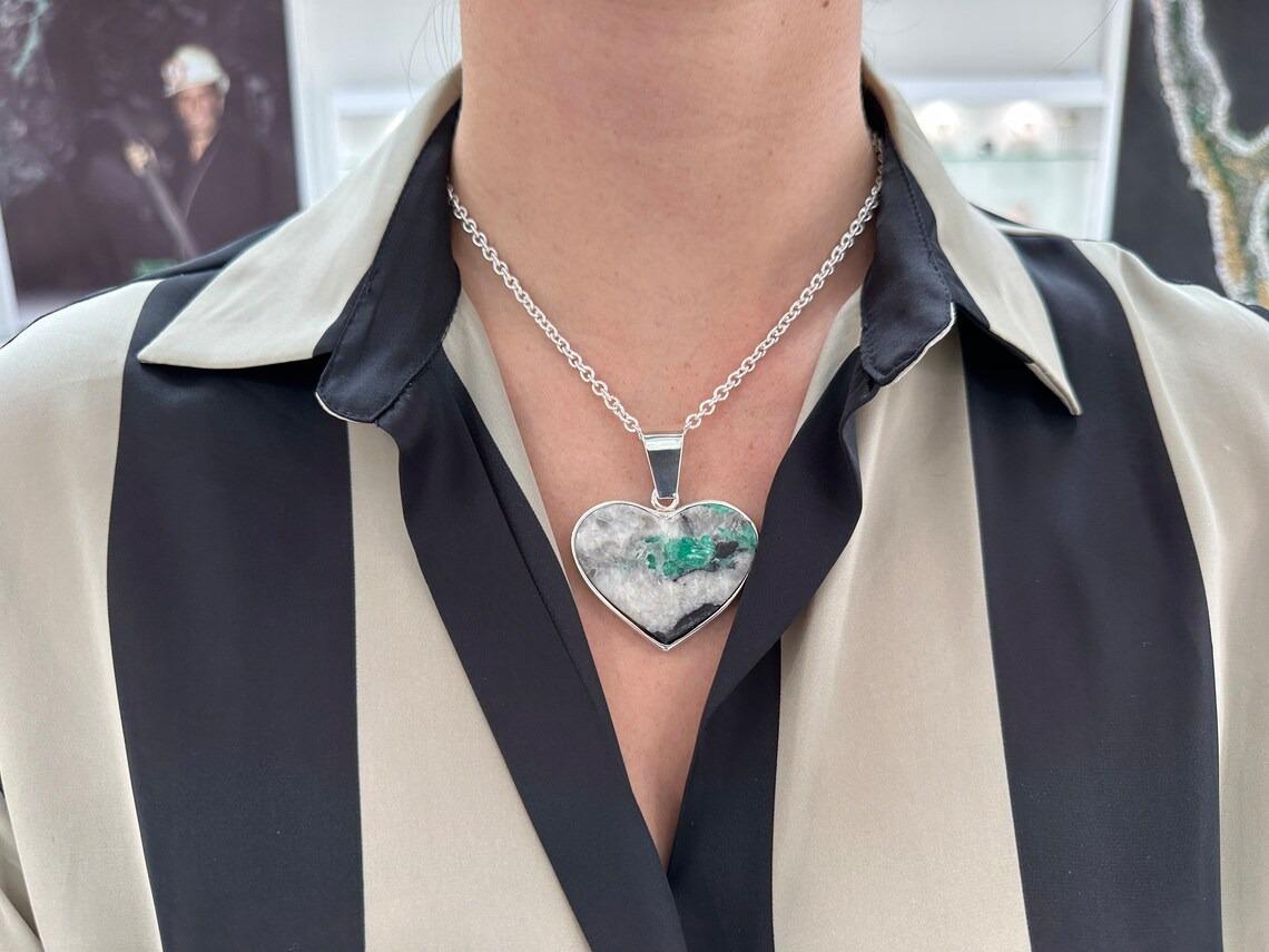 Showcased is a natural, rough emerald embedded in its mother rock, faceted in a heart shape. A stunningly raw, Colombian emerald crystal is bezel set in sterling silver. The emerald showcases a rich green color and is surrounded by white and black