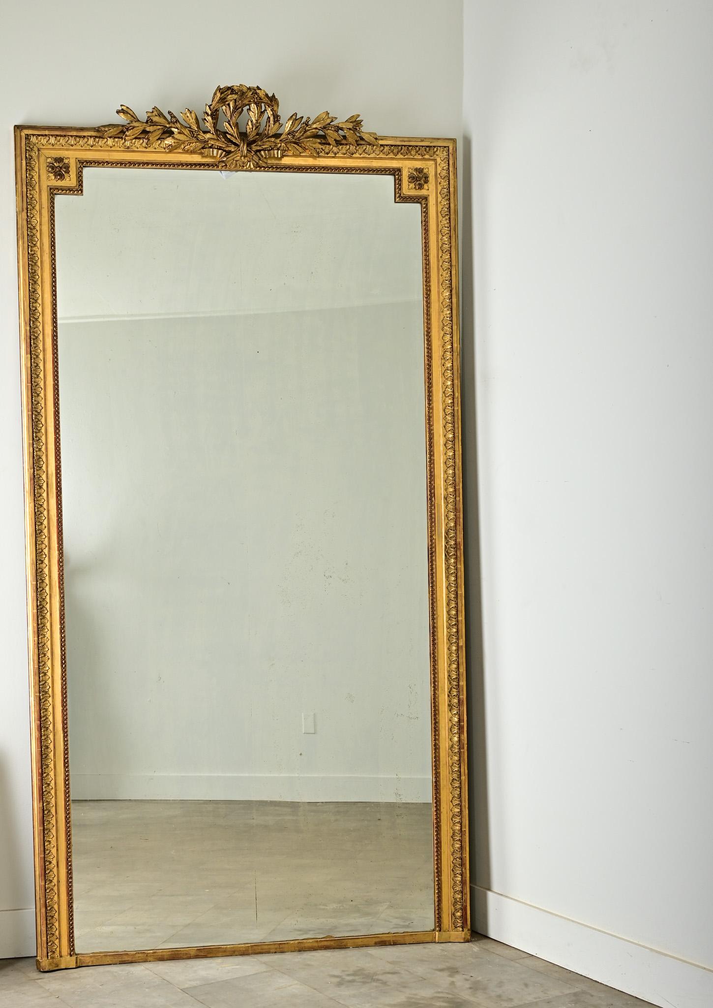 Miroir de cheminée massif de style Louis XVI, avec plaque de miroir d'origine. Le cadre en bois doré est surmonté d'une sculpture représentant deux couronnes de laurier superposées, peut-être un symbole de réussite doublement auspicieux, qui