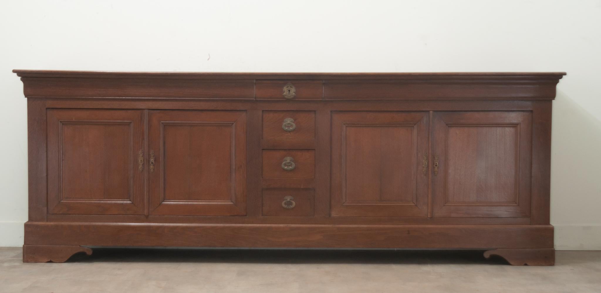 A massive over 9 feet long solid oak Louis Phillipe style French enfilade. The enfilade has a simple plank top. In the center of this case piece you’ll find three true drawers and one faux drawer flanked by two paneled doors. The interior opens with