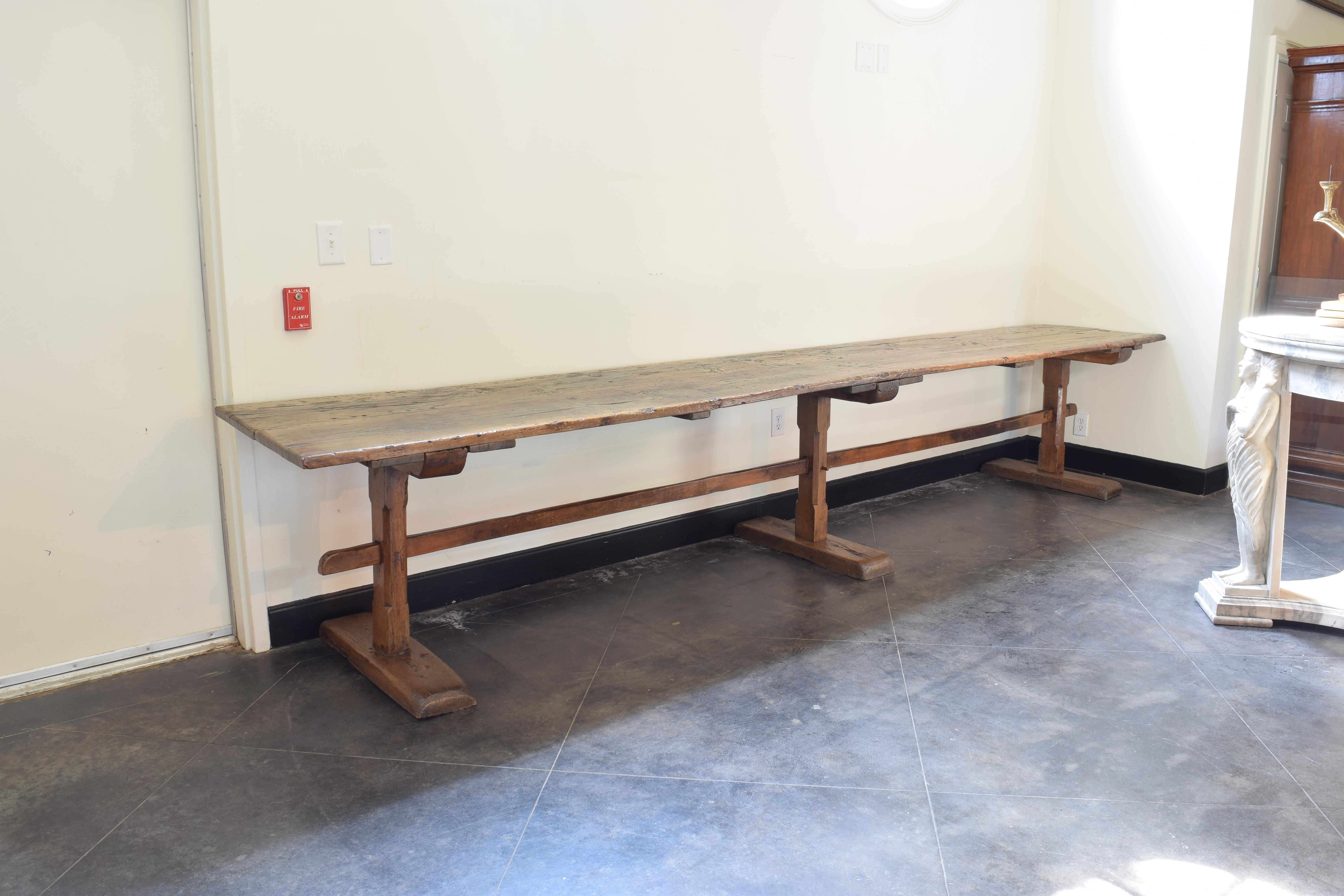 Made for and used in a monastic setting for dining, this table is constructed from two massive wooden planks resting atop three trestles with canted carvings and large bracket feet, the stretcher piercing throught each trestle for stability, in