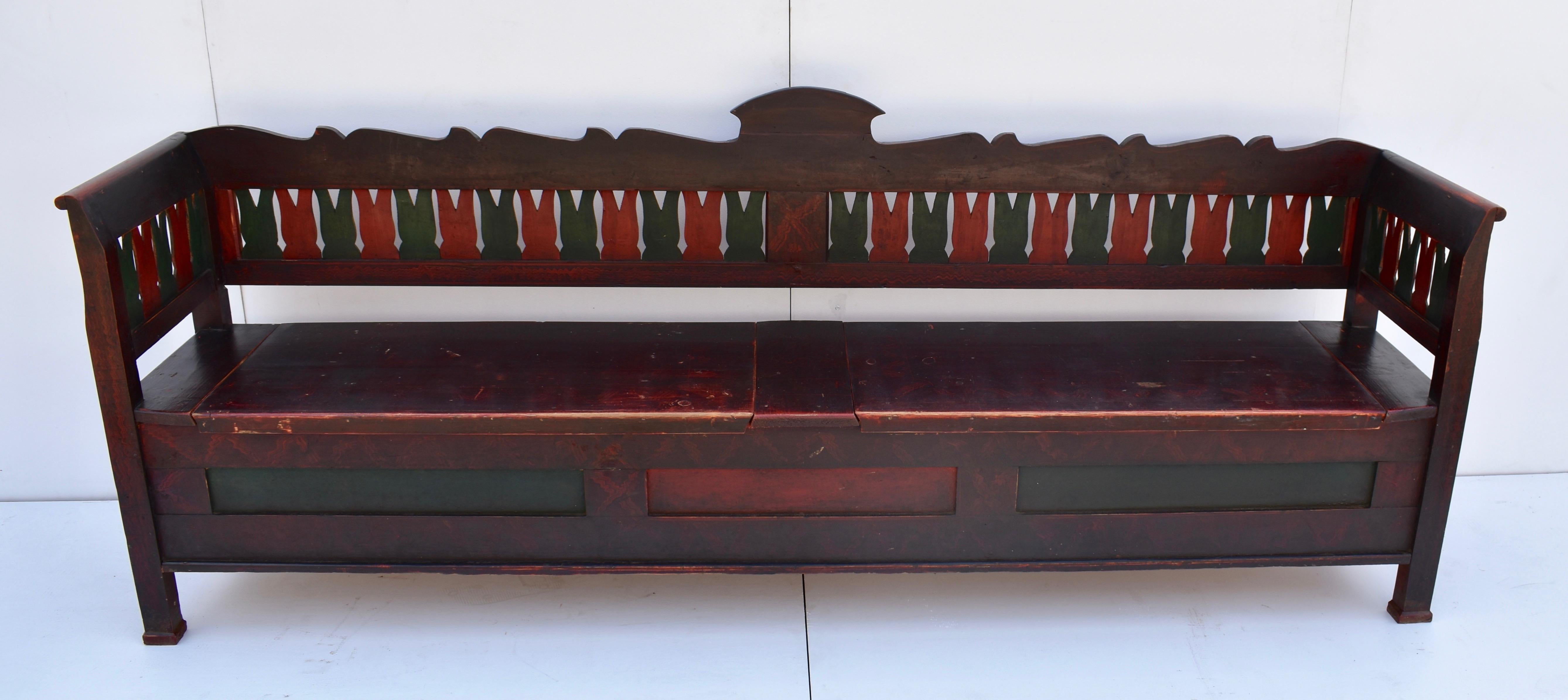 This massive pine storage bench has a nicely scalloped back rail with a crest in the center. The ground is a stylized grain paint in shades of burgundy. Red and green splats alternate around the area between the back and arm rails. The deep storage