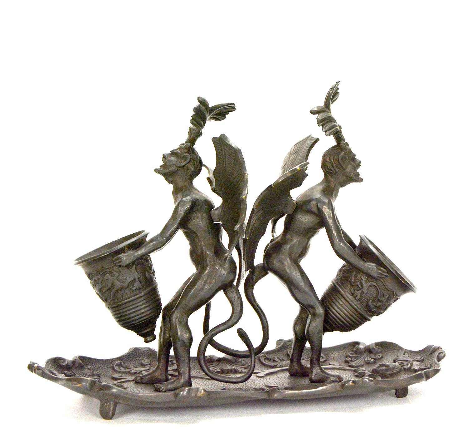 This is a rare massive double Devil figure desk tray match holder. The winged devils are holding a large vase in front of their chest. The vase can be used to store matches. The pair Devils stand on a high relief floral tray, which can be used as
