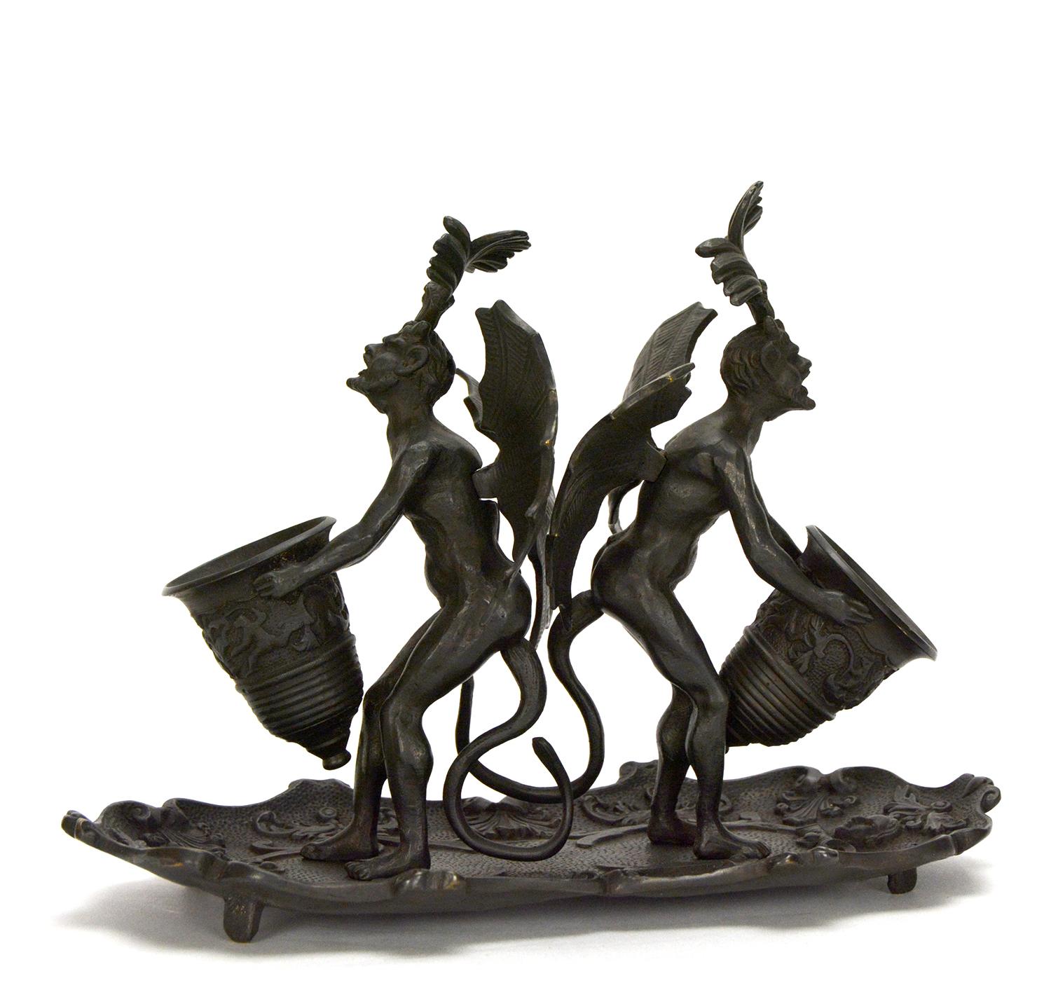 This is a rare massive double Devil figure desk tray match holder. The winged devils are holding a large vase in front of their chest. The vase can be used to store matches. The pair Devils stand on a high relief floral tray, which can be used as
