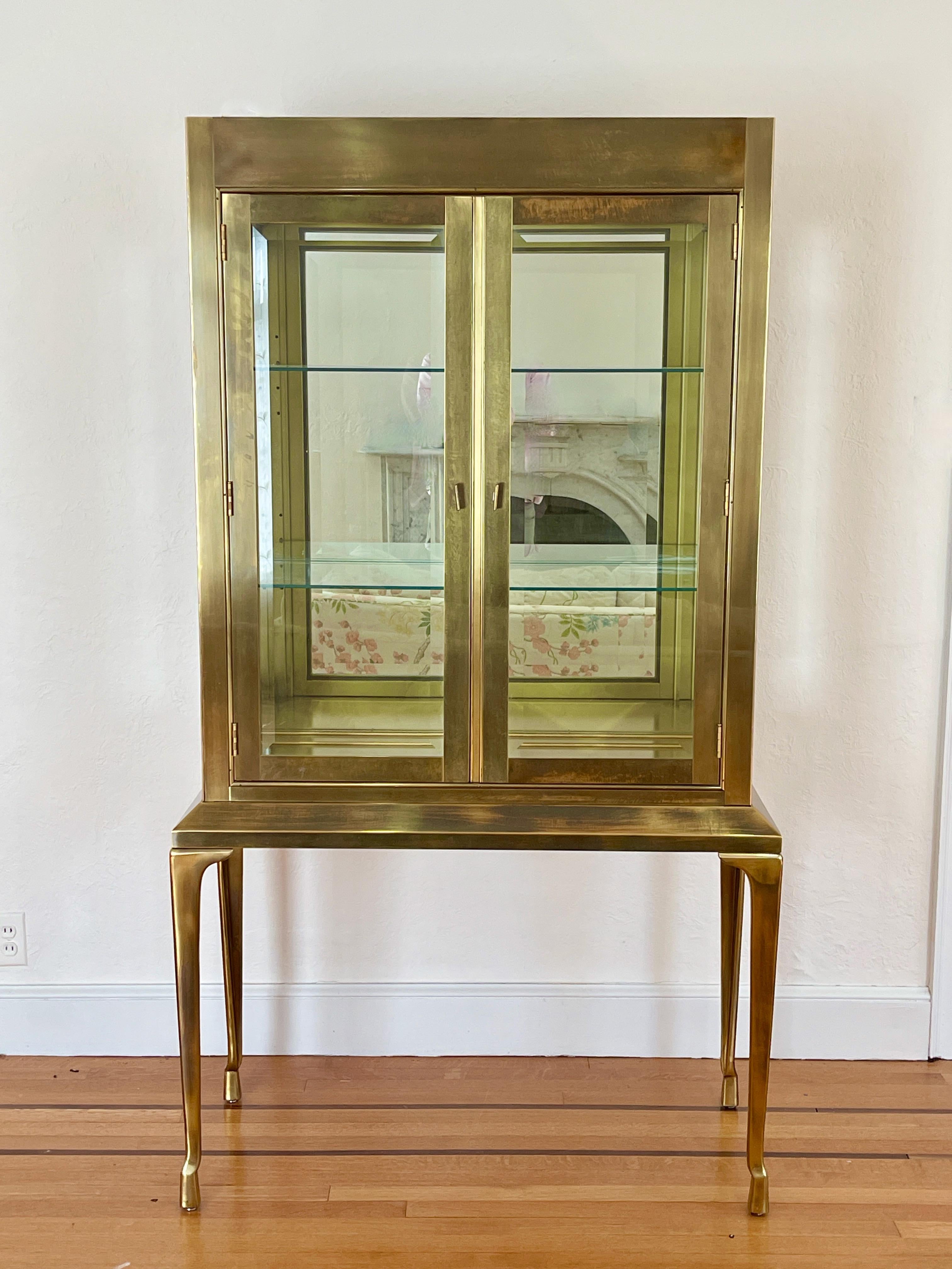 Seldom seen Mastercraft brass display cabinet vitrine showcase on four tall tapering brass Legs 
Interior is illuminated from above by two lightbulbs operated from cord switch.
Backboard mirror.
Two glass shelves with plate grooves.
37.5 x 13 x 33.5