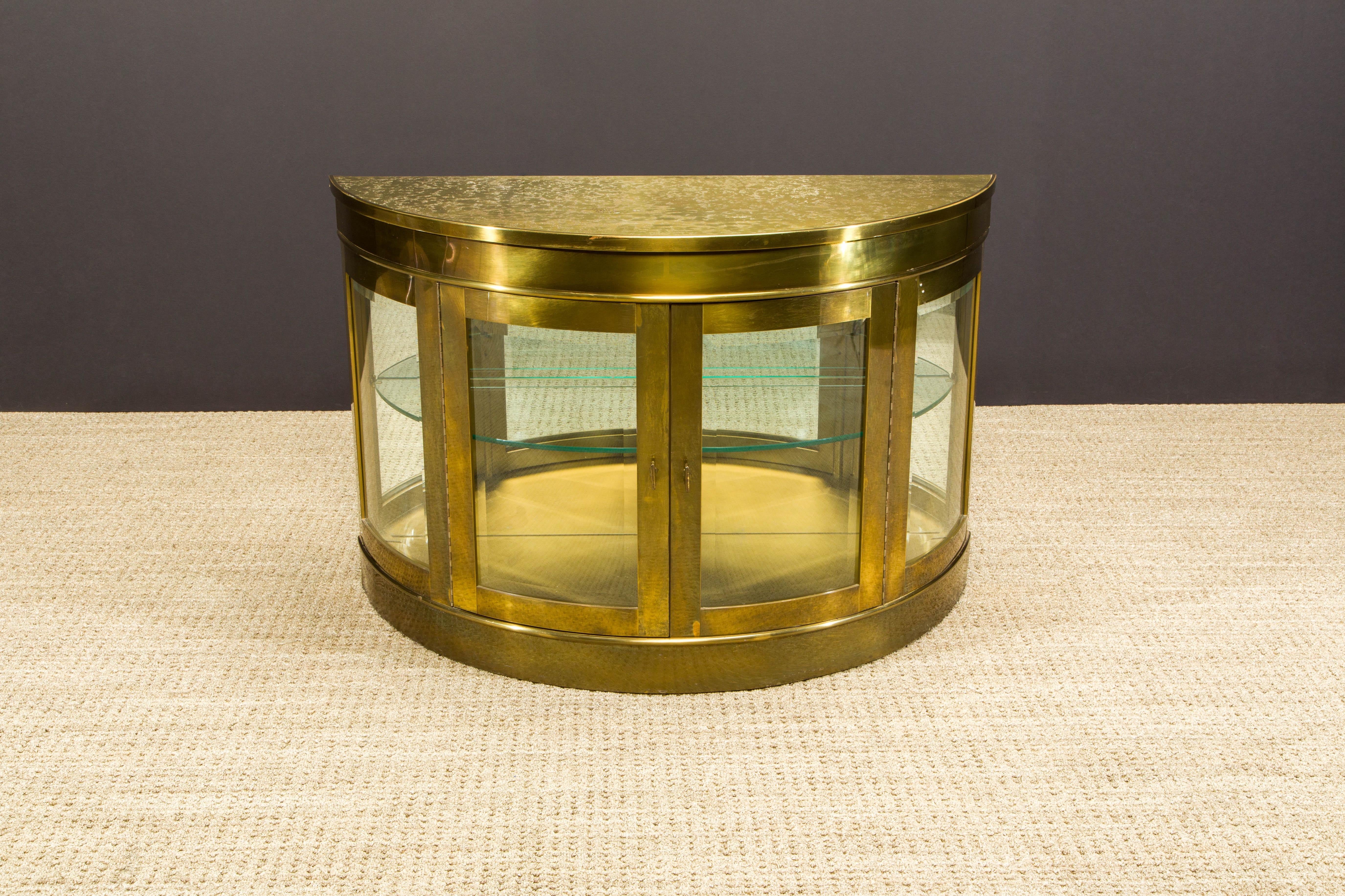 This incredible brass demilune display cabinet, produced by Mastercraft in the 1970s, features a mirrored and illuminated interior, exquisite textured top, a pair of glass doors and interior glass shelf. 

What makes this amazing design even more