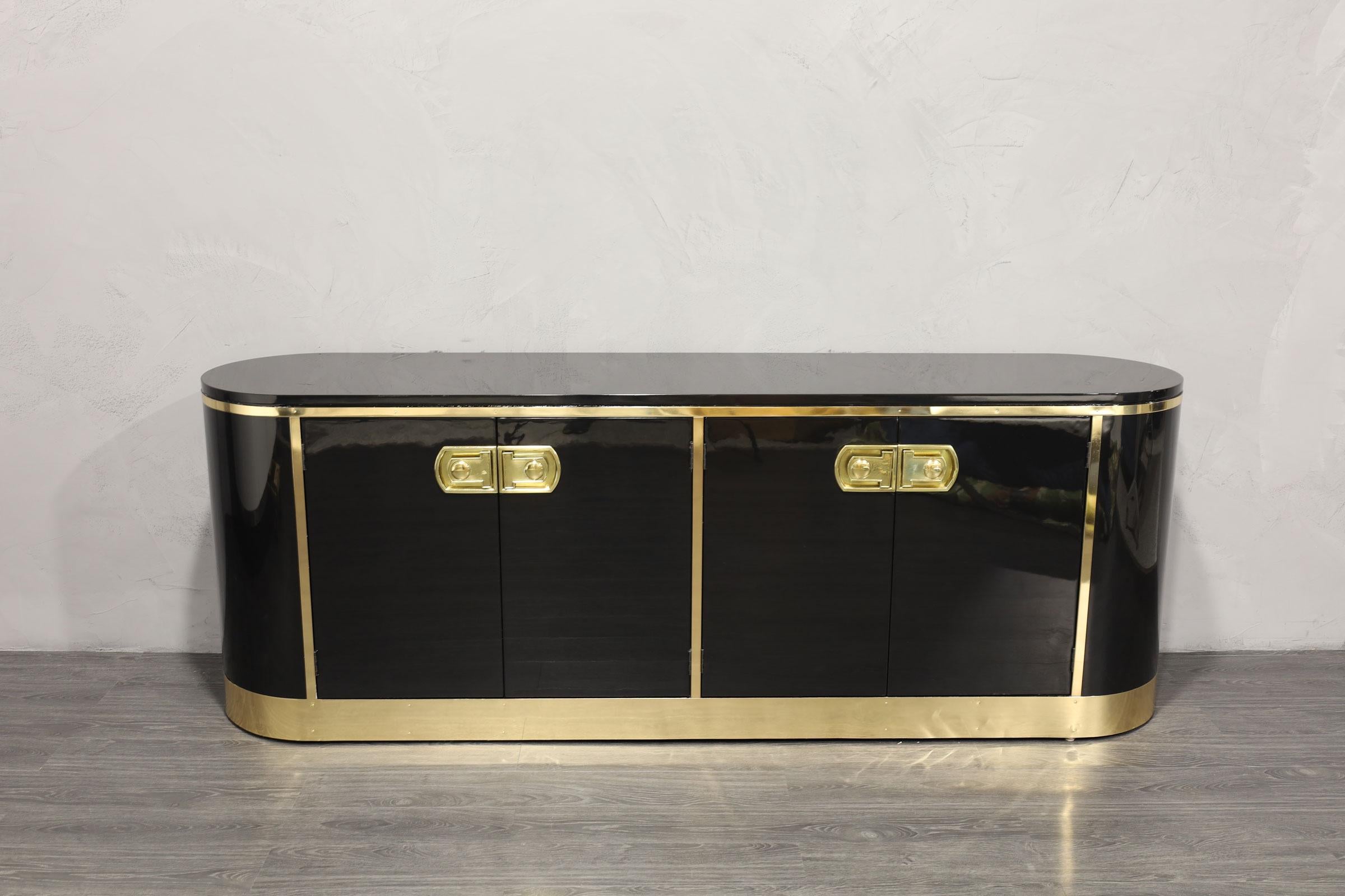 A beautifully restored sideboard by Mastercraft. It is pill shaped and an iconic design. This is a high gloss black lacquer with shelving on one side and four drawers on the other side. Brass trim that has been expertly polished and sealed so it