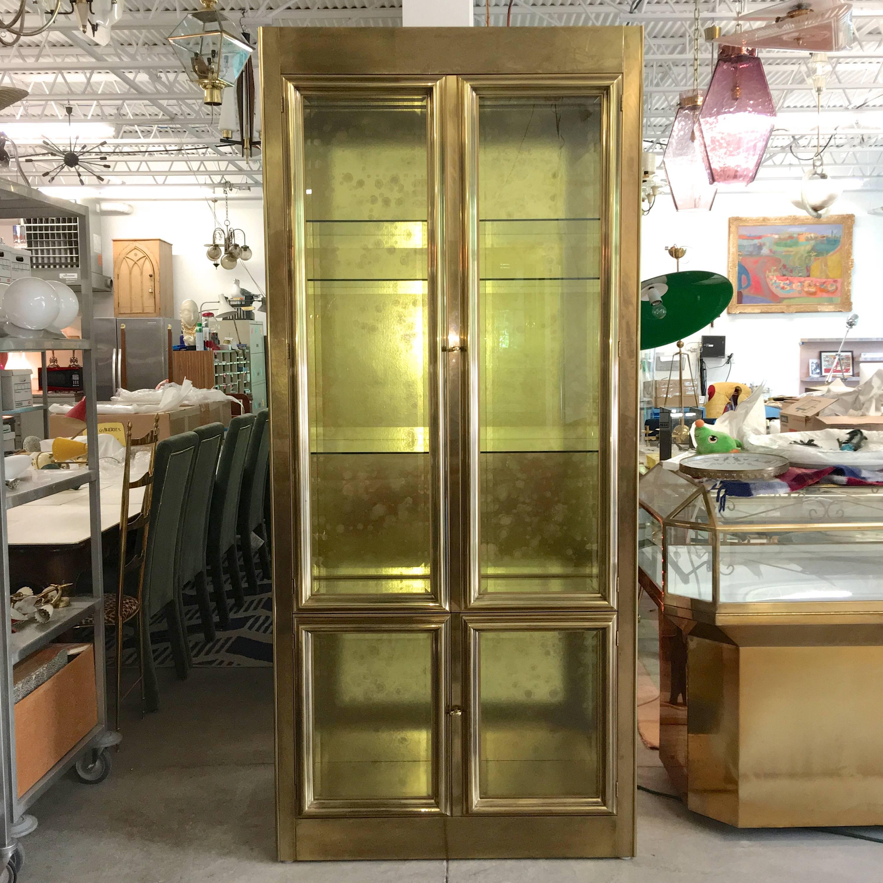 Mastercraft brass and glass four door vitrine with dimmable overhead lights. Large double doors over small double doors, both with clear beveled glass. Beveled glass window sidelights. Gold foiled backboard. Three adjustable shelves with etched