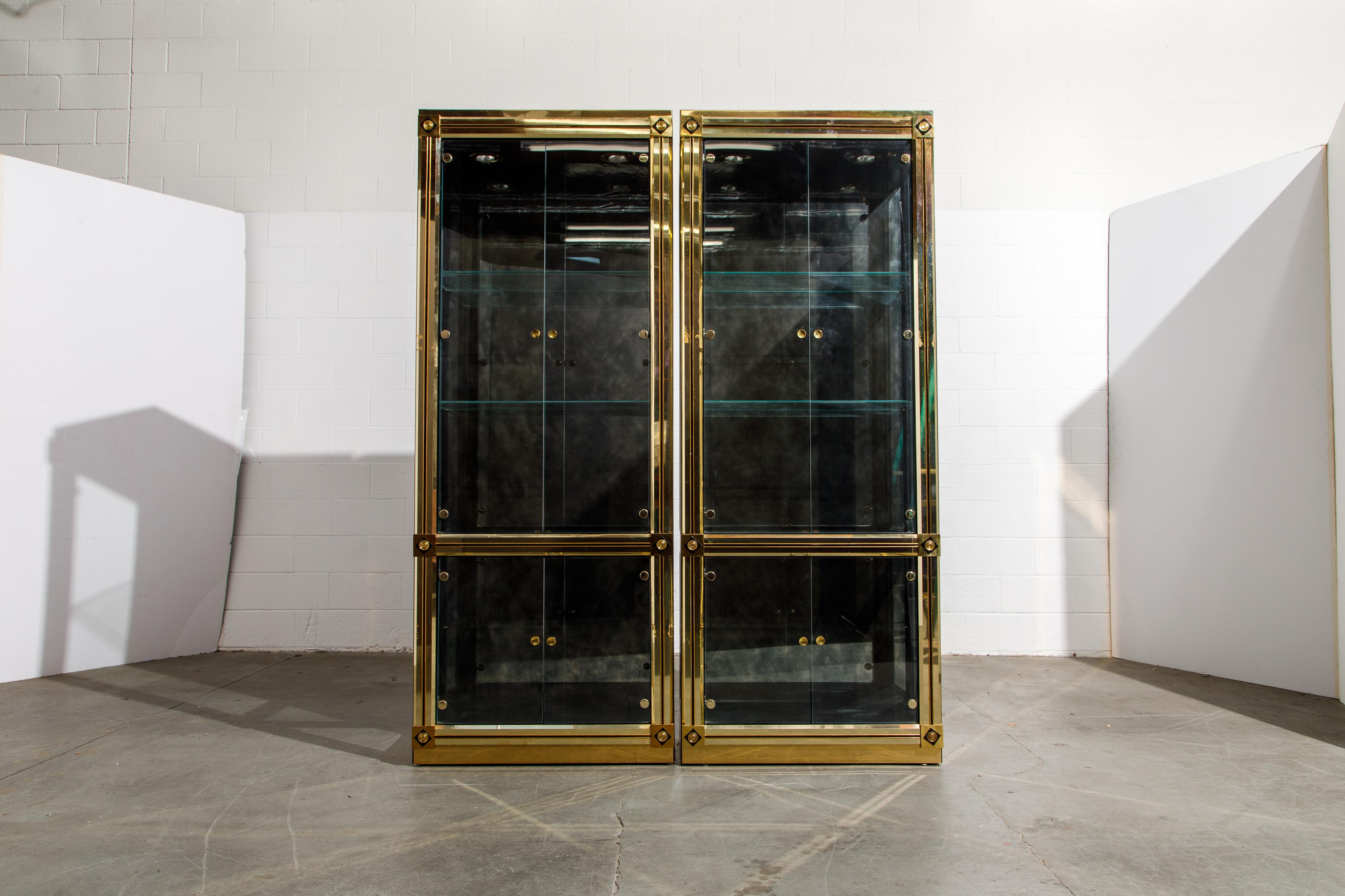 A beautiful pair of illuminated brass vitrines by Mastercraft, circa 1980 or earlier. With ebonized accents and four glass doors, these display cabinets have antique mirrored backs and adjustable clear glass shelves, each with two dimmable recessed