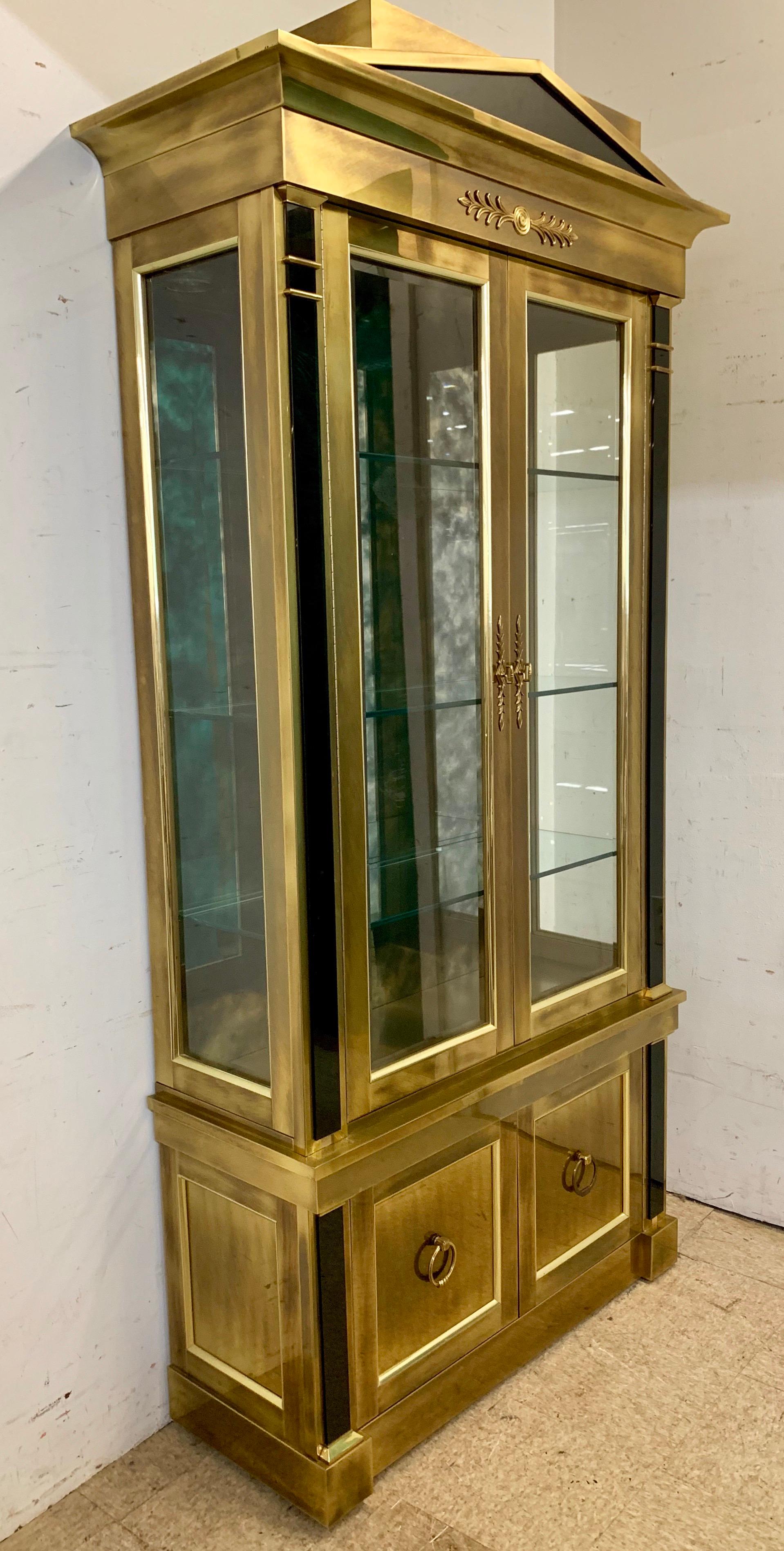Stunning Mastercraft display cabinet made of brass with black glass accents. Glass doors open to glass shelves with mirrored background and is lighted from above. Bottom brass doors open to more storage. Top pediment and architectural detail give it