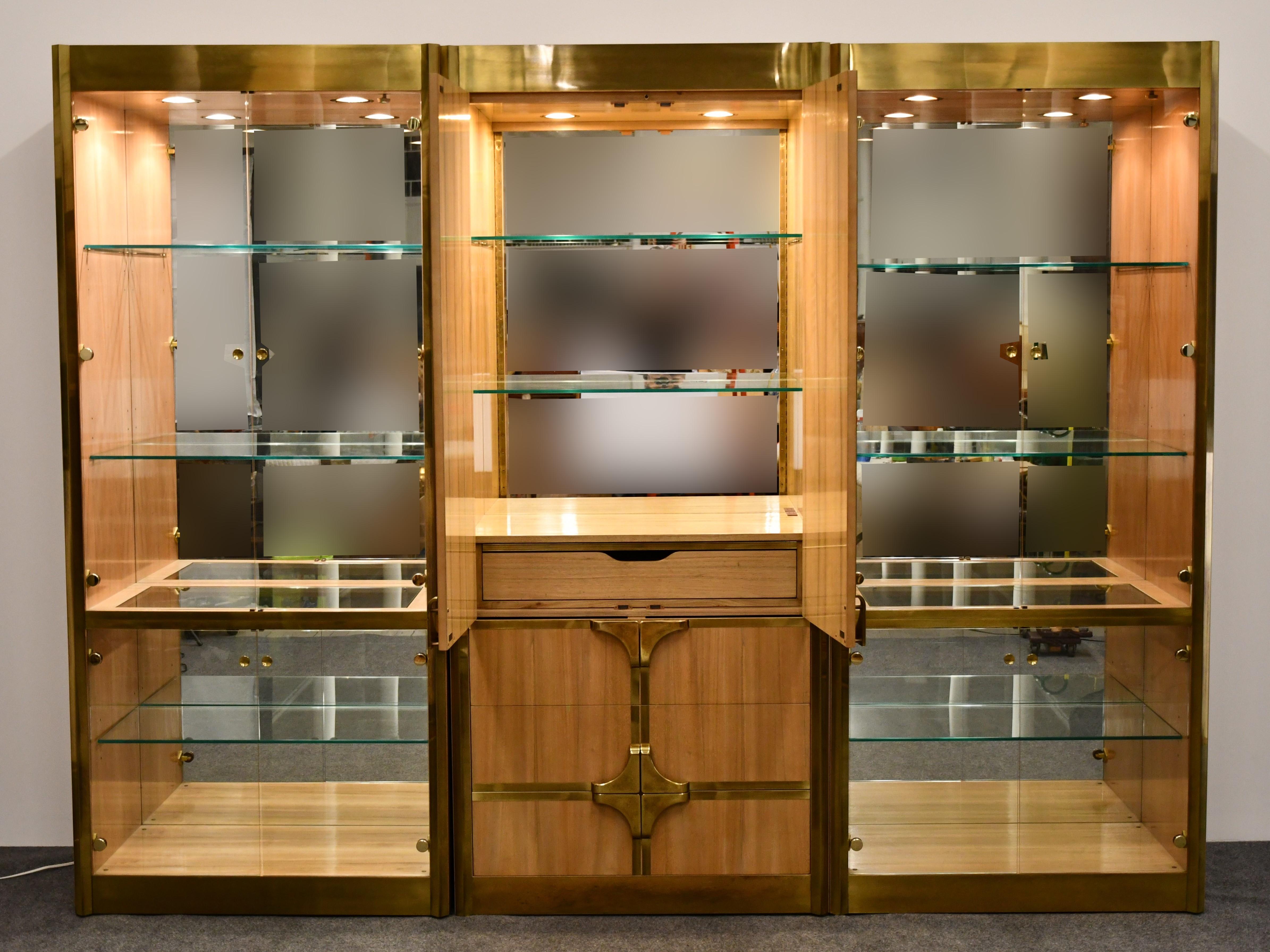 A magnificent three-piece Mastercraft brass and bleached walnut vitrine. This wall cabinet has six interior dimmable downlights, four drawers, and ten glass shelves. Very good vintage condition with age appropriate wear. Several scratches to the