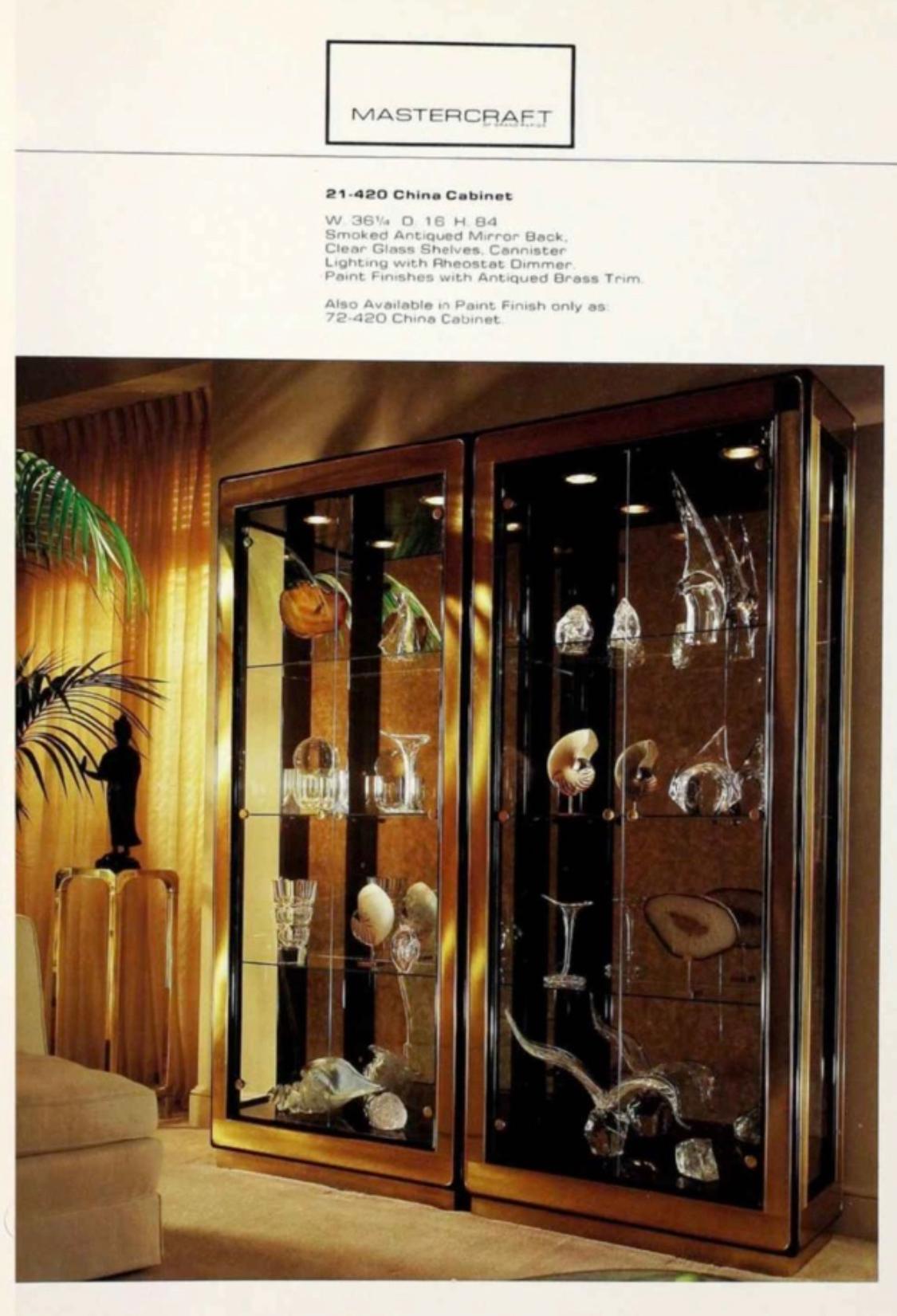 Mastercraft model 21-420 china cabinet vitrine. Clad in antiqued brass with black lacquer trim. Backwall is smoked antiqued mirror.  Three tempered clear glass shelves with plate grooves.  Illuminated by two cannister lights in ceiling controlled by