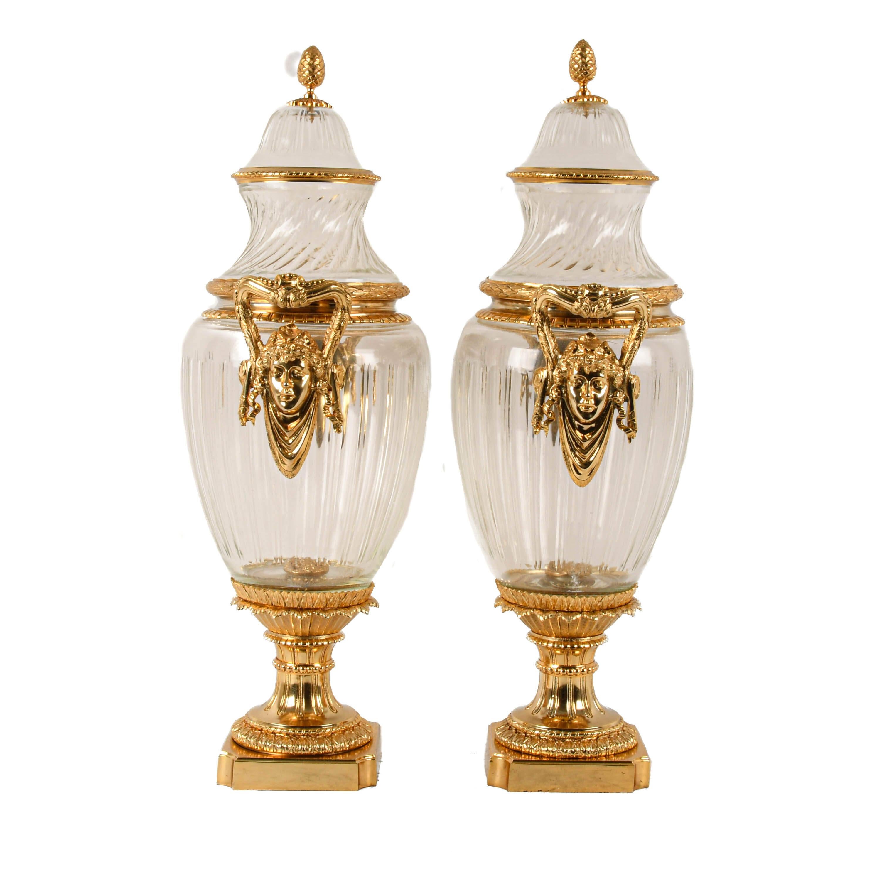 Here is pure grandeur in a matched pair. These Baccarat-style gilt bronze and crystal urns are a masterclass in luxury and refinement. The hand-cut crystal bodies shimmer with clarity, each vertical flute capturing light like a prism. Framed by