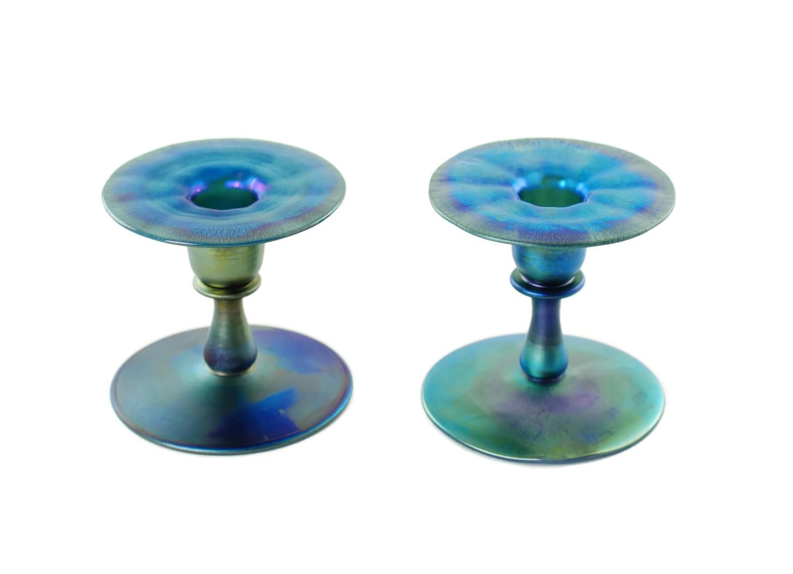 This matched pair of handcrafted Louis Comfort Tiffany single light candleholders have been made in the company's striking Blue Favrile art glass. The pieces have round pedestal bases with short baluster form stems. The stems are topped by candle