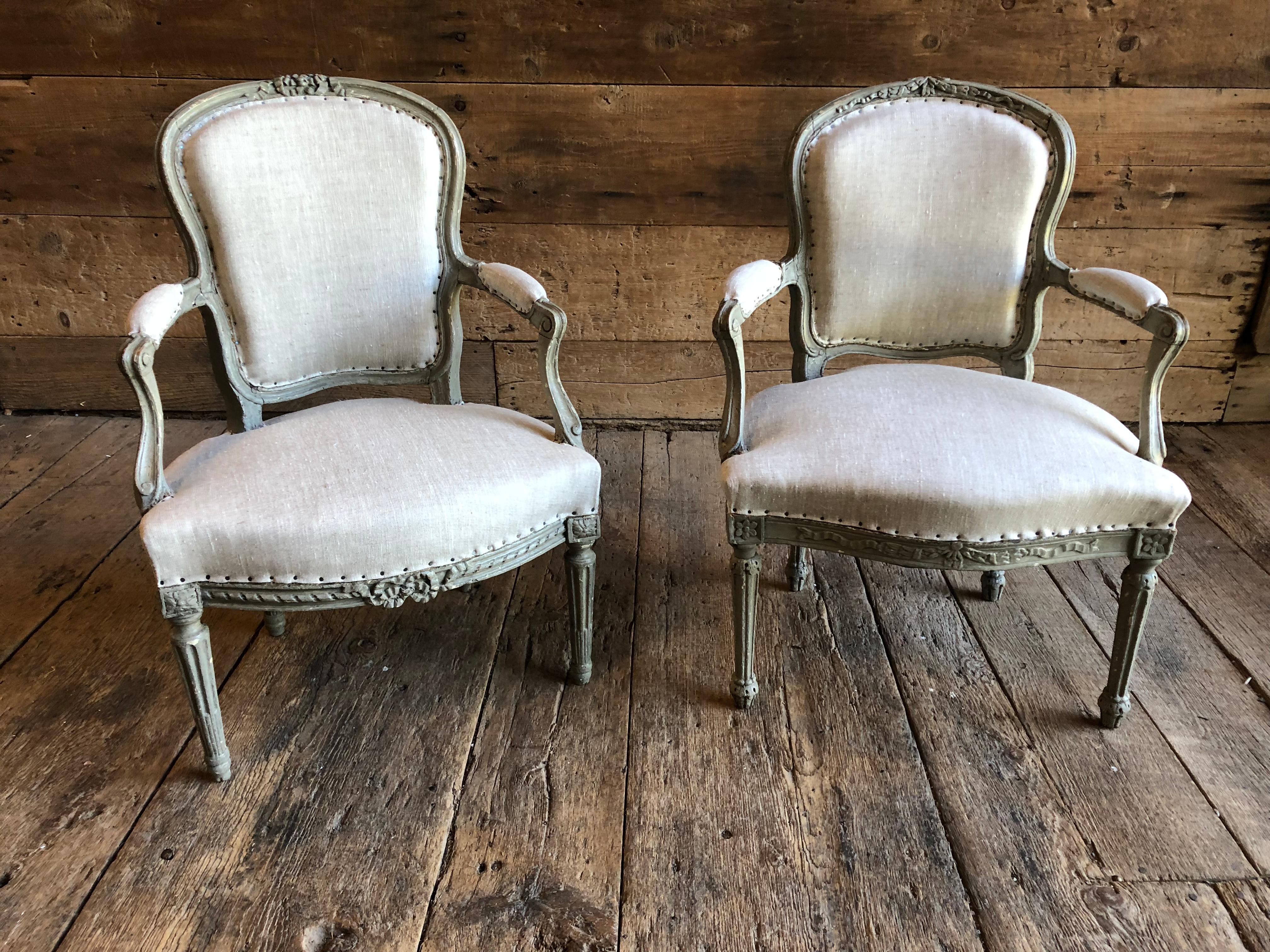 A matched companion pair of transitional Louis XVI period fauteuils in old grey painted finish, the arms showing elements of the early Louis XV period with subtle curves and scrolls, the back and seat rail with floral and ribbon carvings, both on