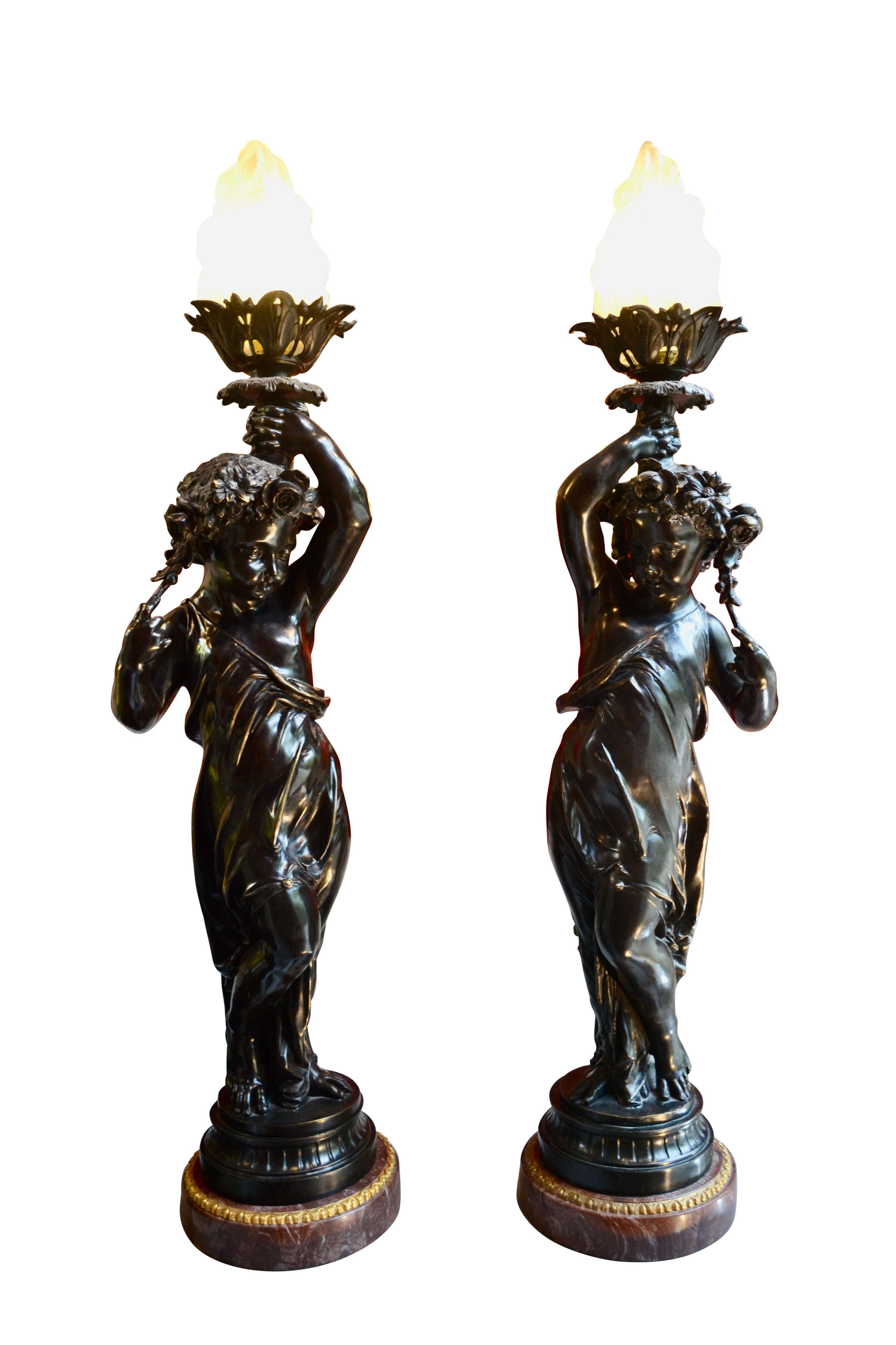 A beautifully cast matching pair of patinated bronze figural torcheres in the manner of Clodion modeled as classically draped young girls with garlands of flowers in their hair and holding aloft a flame torch. Set on a round marble base with gilt