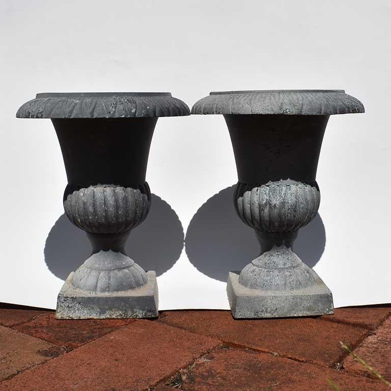 Matched pair of cast iron urn planters with heavy patina on their pale grey finish. Some pitting to the iron of interior bottoms. Dimensions: 9.5