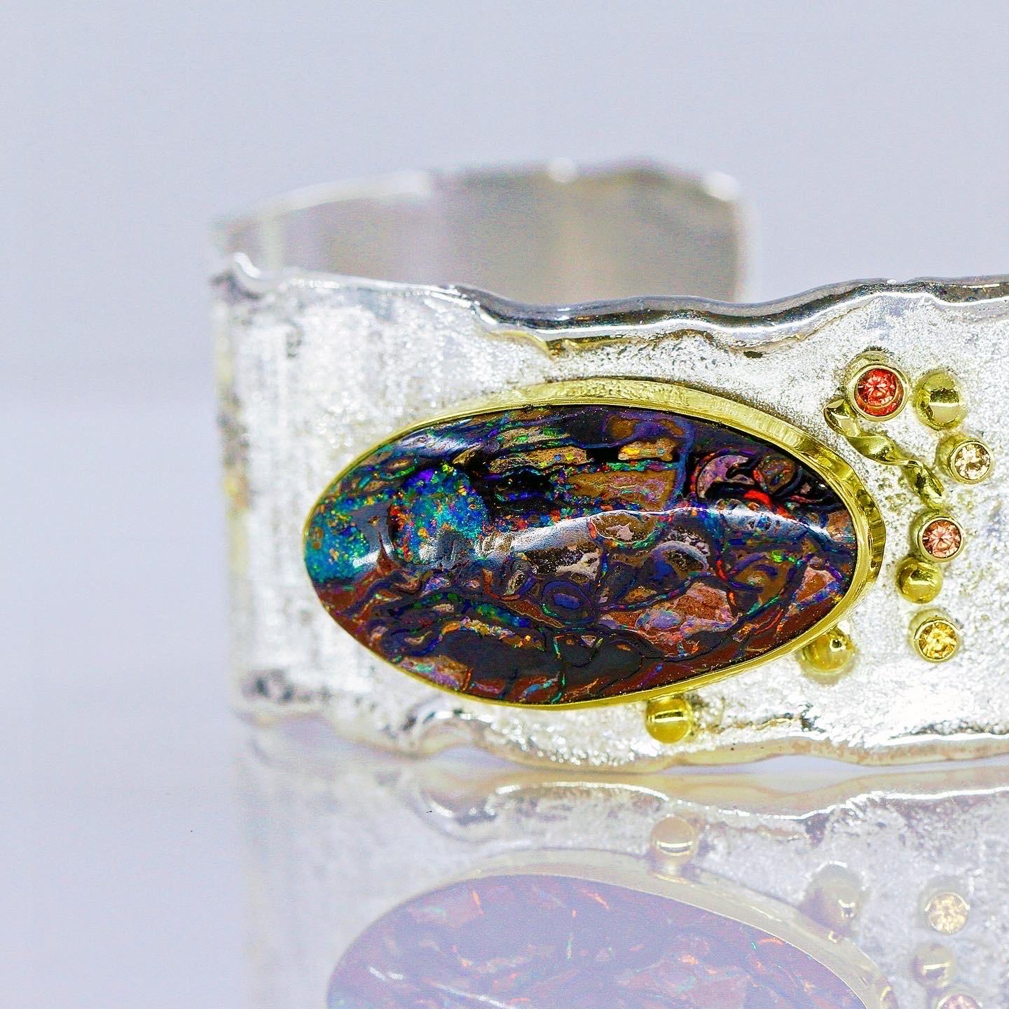 Une opale Matrix remplie de toutes les couleurs que l'opale peut offrir.  L'opale est enveloppée dans de l'or 22k, des saphirs d'appoint complètent et de la poussière d'or est saupoudrée sur le bracelet manchette en argent sterling.  Le bracelet
