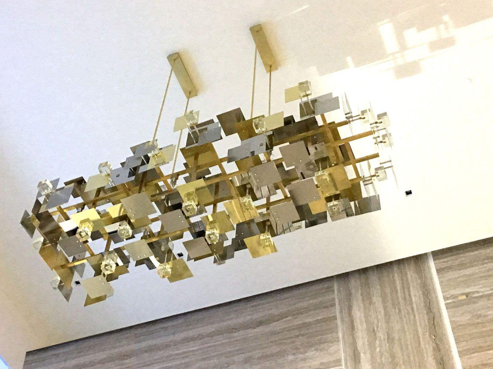 Mixed metals, fused and cast glass components combined to create a strikingly geometric, warmly glowing chandelier.
Brushed, polished brass and stainless steel, fused glass panels and clear cast glass cubes attached to central brass