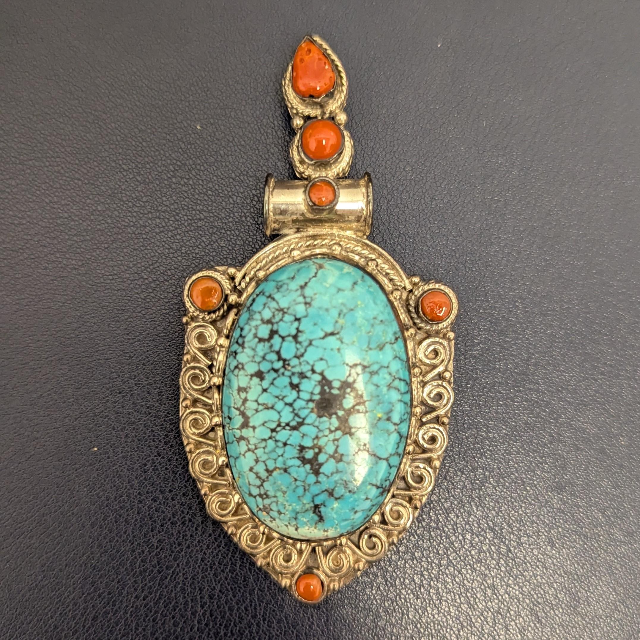 This piece can be worn as either a pendant or brooch. It features a center bright blue turquoise, with black matrix throughout and slight fading of the color at the ends, accented with bright orangy red, various shaped corals, set in sterling silver.
