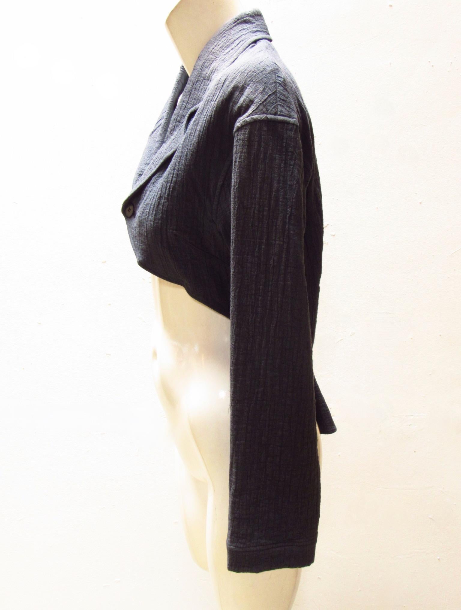Cette veste cropped en coton du millésime Matsuda est de couleur pétrole, le tissu a une texture de gaze lourde. Les revers se ferment à l'aide d'un bouton en bois à l'avant, au niveau de la partie centrale du vêtement. Le tissu plus long dans le