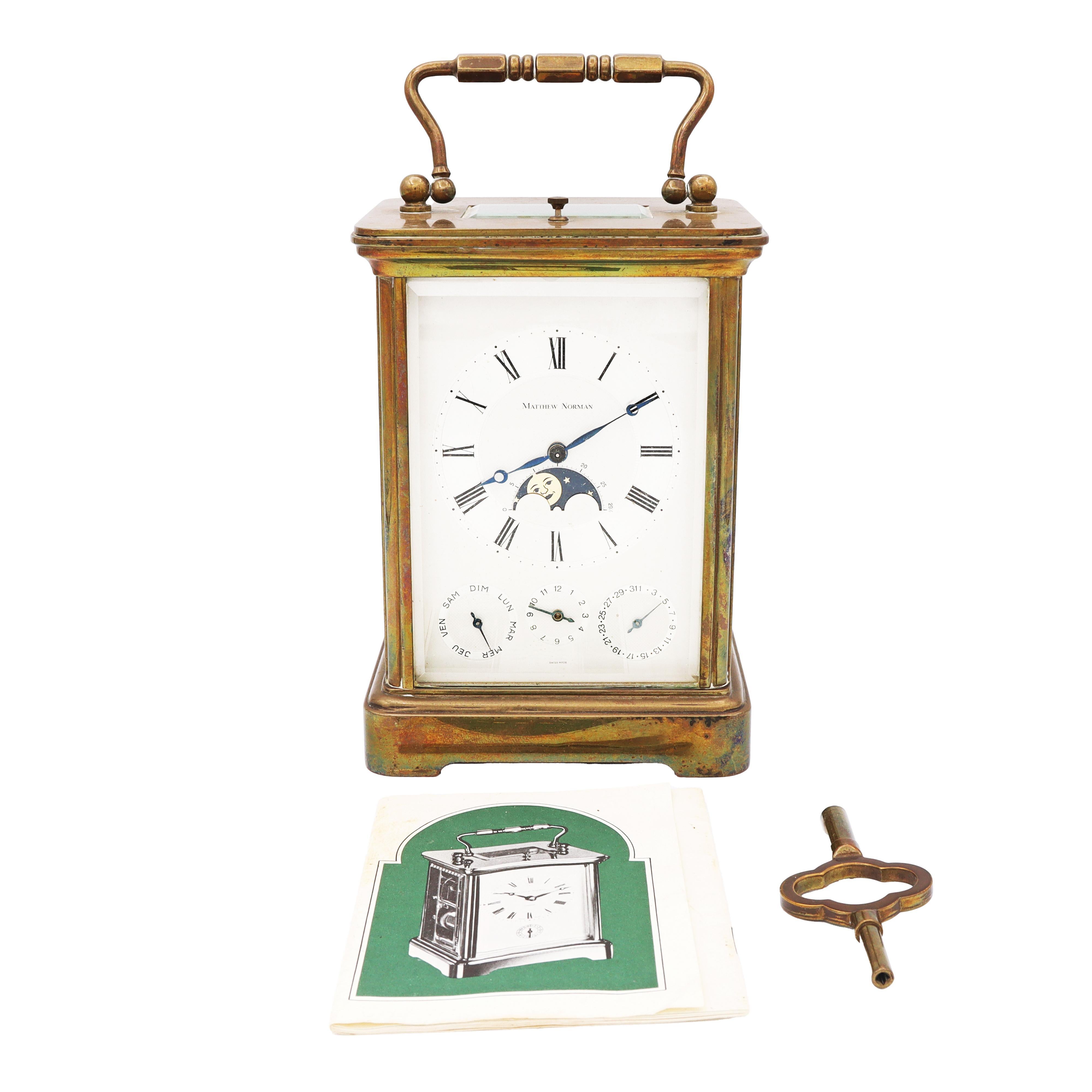 This elegant Matthew Norman brass-cased carriage clock is a beautifully crafted timepiece that showcases both classic charm and intricate functionality. Housed in a patinated brass case with beveled glass panels and a decorative carrying handle,